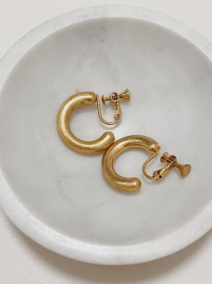 A pair of Zora brushed gold clip-on hoop earrings with adjustable screw back clasps lying on a minimal white marble dish.