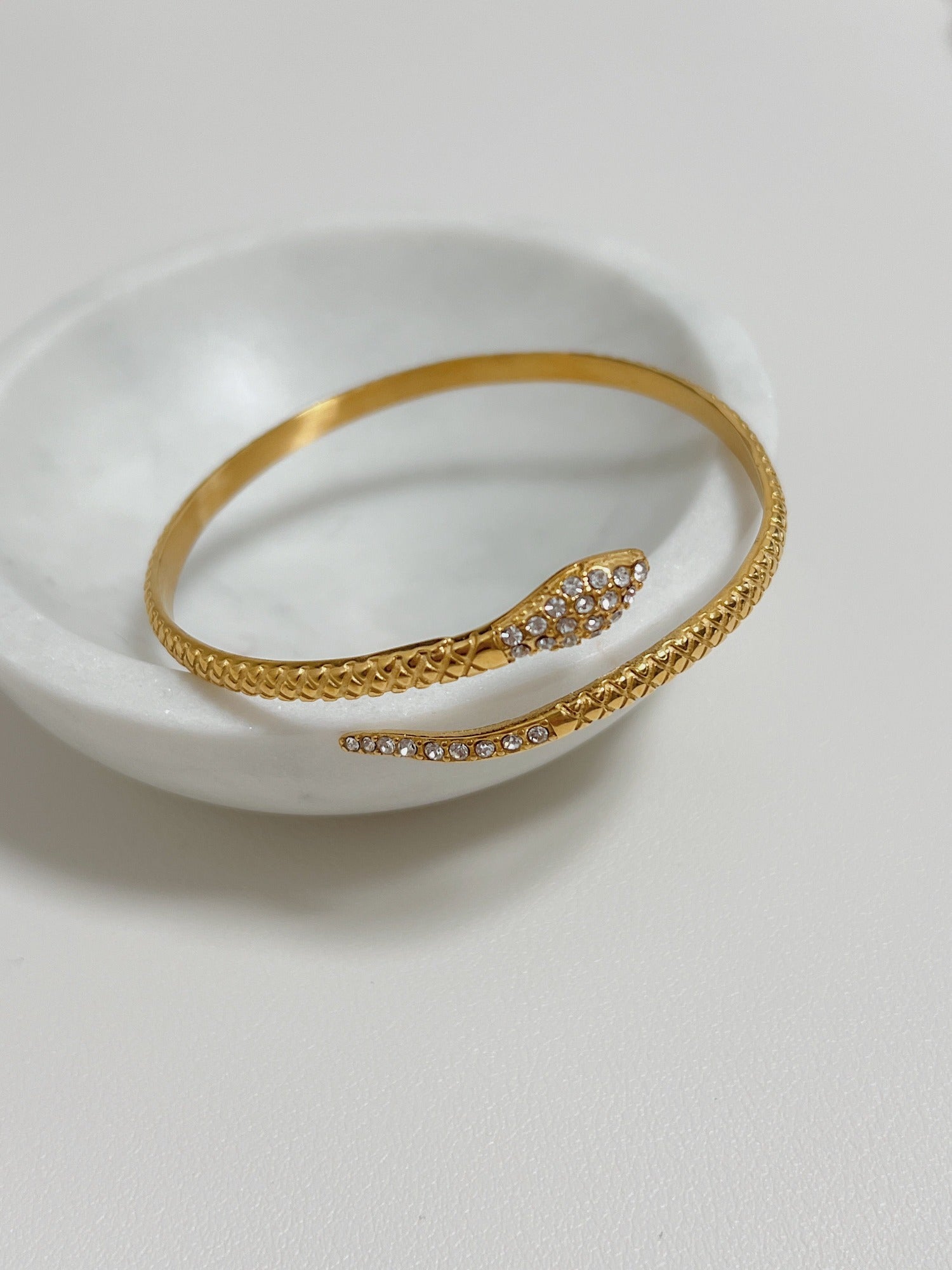 18K gold Isa Serpent Bangle resting on a white marble bowl highlighting the 5A CZ pavé head and scale details.