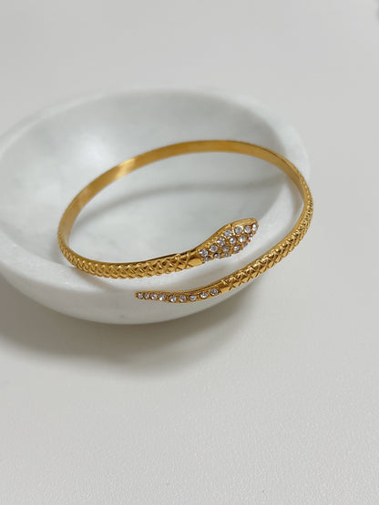 18K gold Isa Serpent Bangle resting on a white marble bowl highlighting the 5A CZ pavé head and scale details.