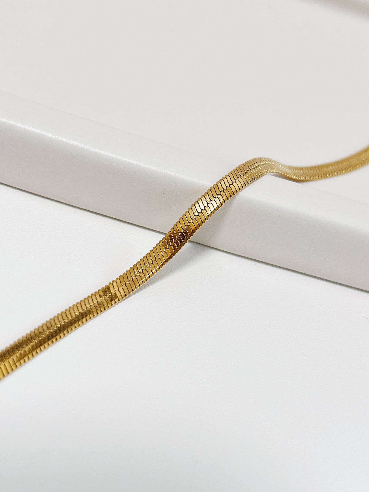 Macro close-up shot of the Bec 18K gold PVD herringbone chain draping smoothly over a curved white porcelain ledge, showing the detailed micro-links