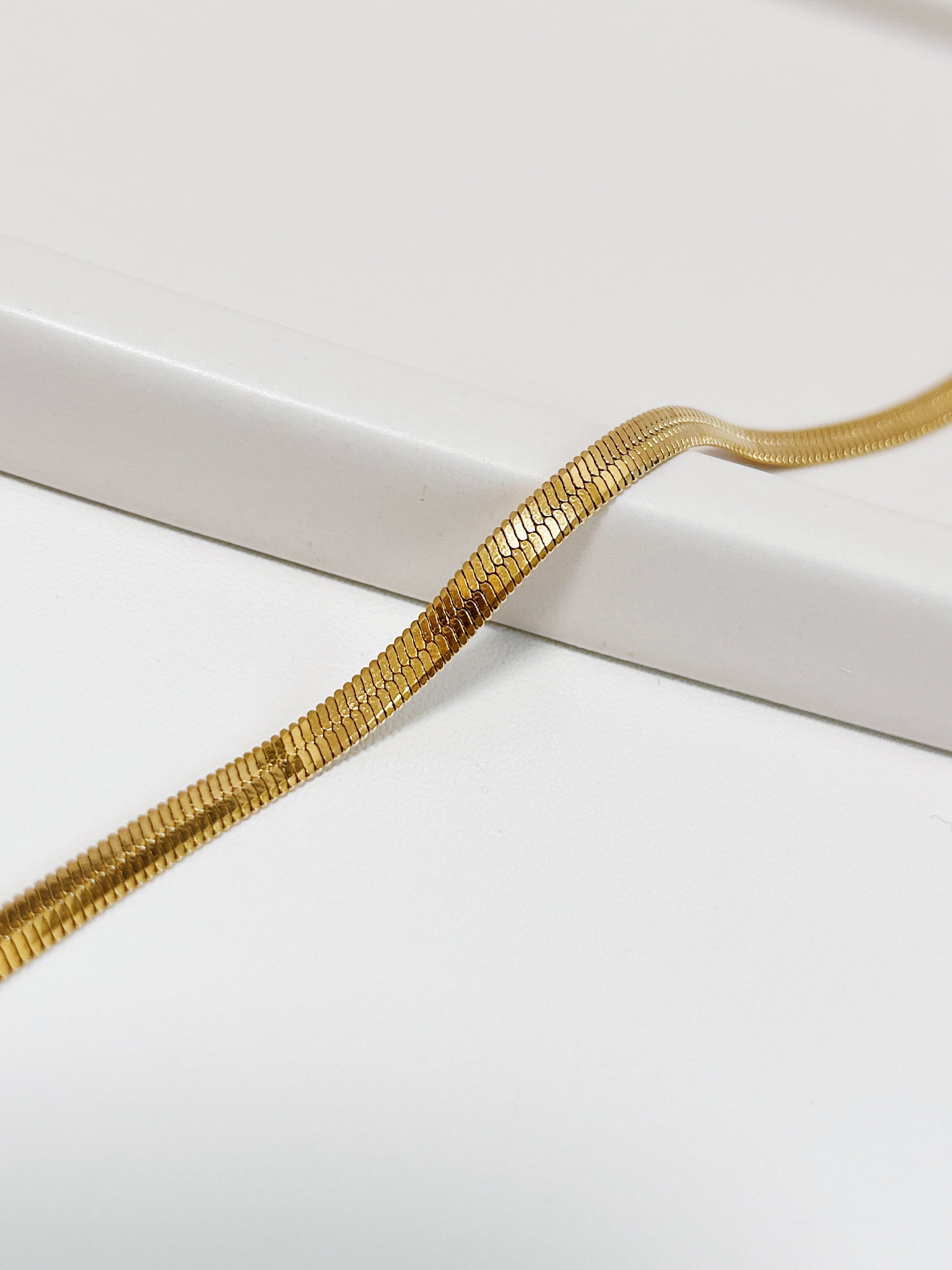 Macro close-up shot of the Bec 18K gold PVD herringbone chain draping smoothly over a curved white porcelain ledge, showing the detailed micro-links