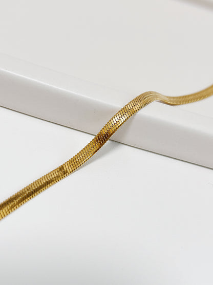 Macro close-up shot of the Bec 18K gold PVD herringbone chain draping smoothly over a curved white porcelain ledge, showing the detailed micro-links