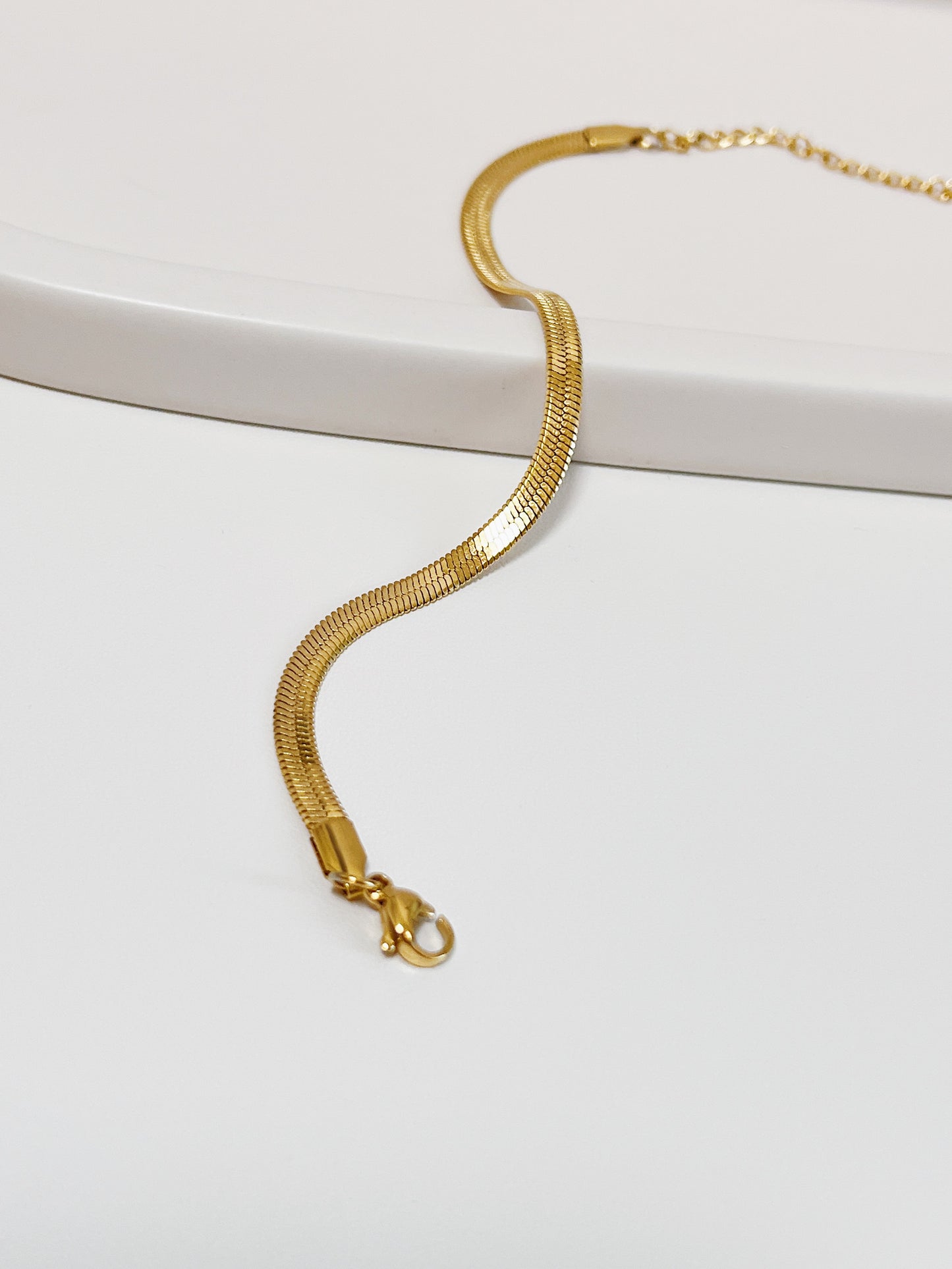 High-angle studio view of the Bec gold flat-woven snake chain arranged in an elegant s-curve across a matte white ceramic tray, showcasing its flexible, fluid drape