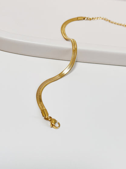High-angle studio view of the Bec gold flat-woven snake chain arranged in an elegant s-curve across a matte white ceramic tray, showcasing its flexible, fluid drape