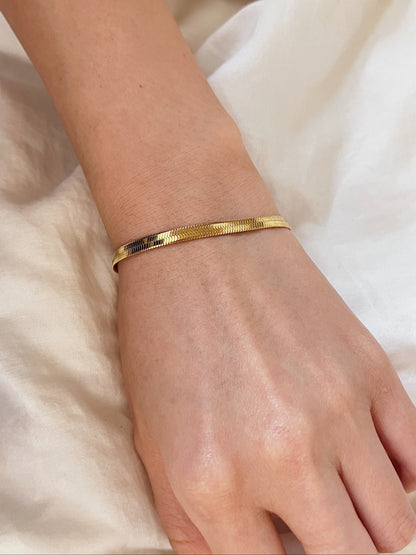 Mid-range view of the Bec gold flat herringbone bracelet styled solo on a model's wrist against a textured cream linen background