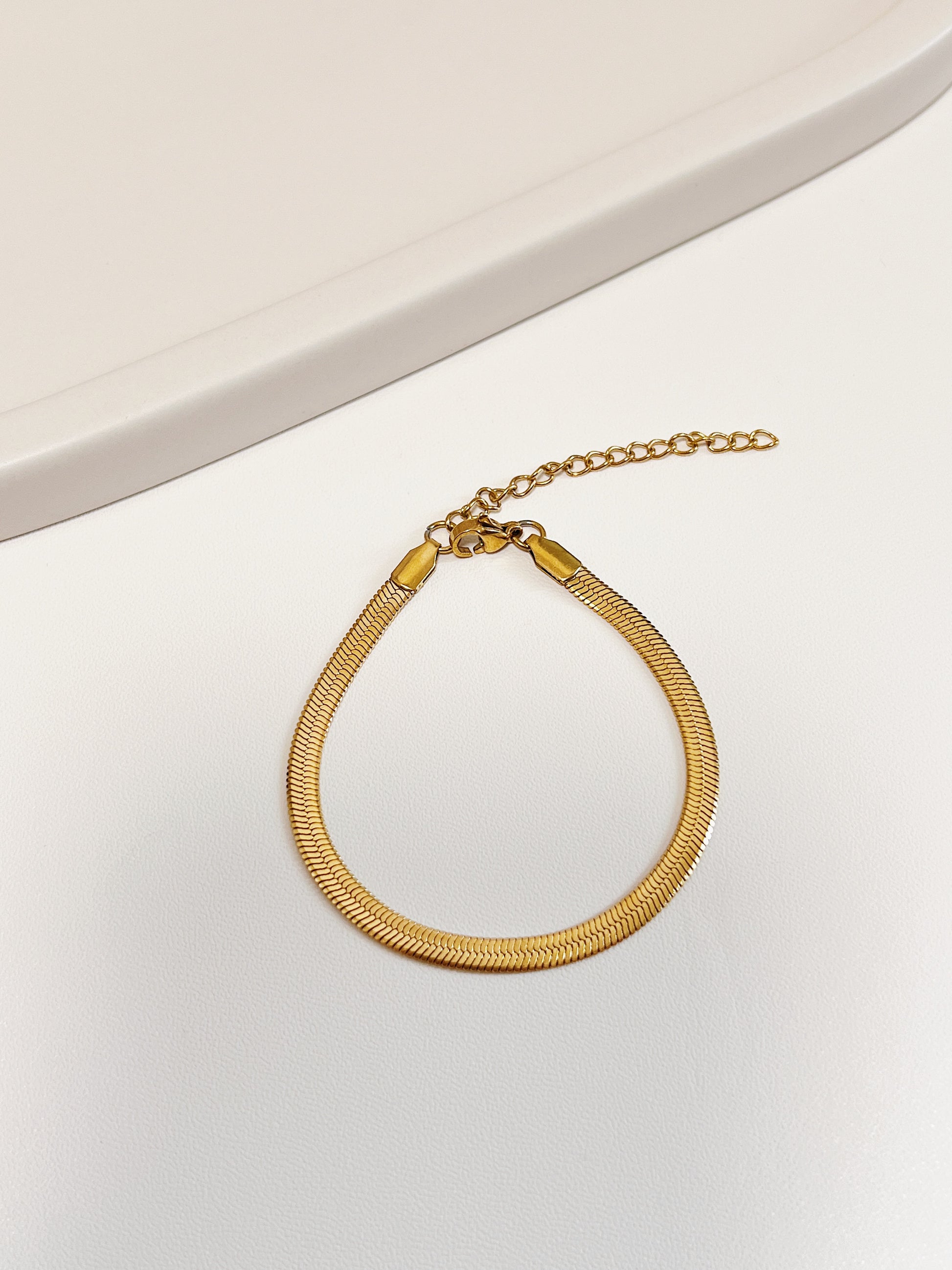 Overhead product view of the Bec gold snake chain bracelet resting in a clean loop on a minimalist white display surface, highlighting the secure lobster clasp and adjustable extension chain