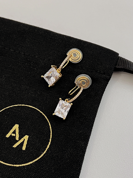 Pair of gold Candice emerald cut drop clip-on earrings with pavé bars resting on black pouch background