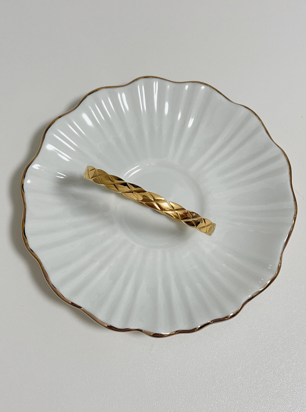 The Corin gold quilted hinged bangle displayed flat inside a delicate, gold-rimmed fluted porcelain dish, highlighting the geometric cross-hatched pattern.