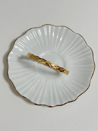 The Corin gold quilted hinged bangle displayed flat inside a delicate, gold-rimmed fluted porcelain dish, highlighting the geometric cross-hatched pattern.