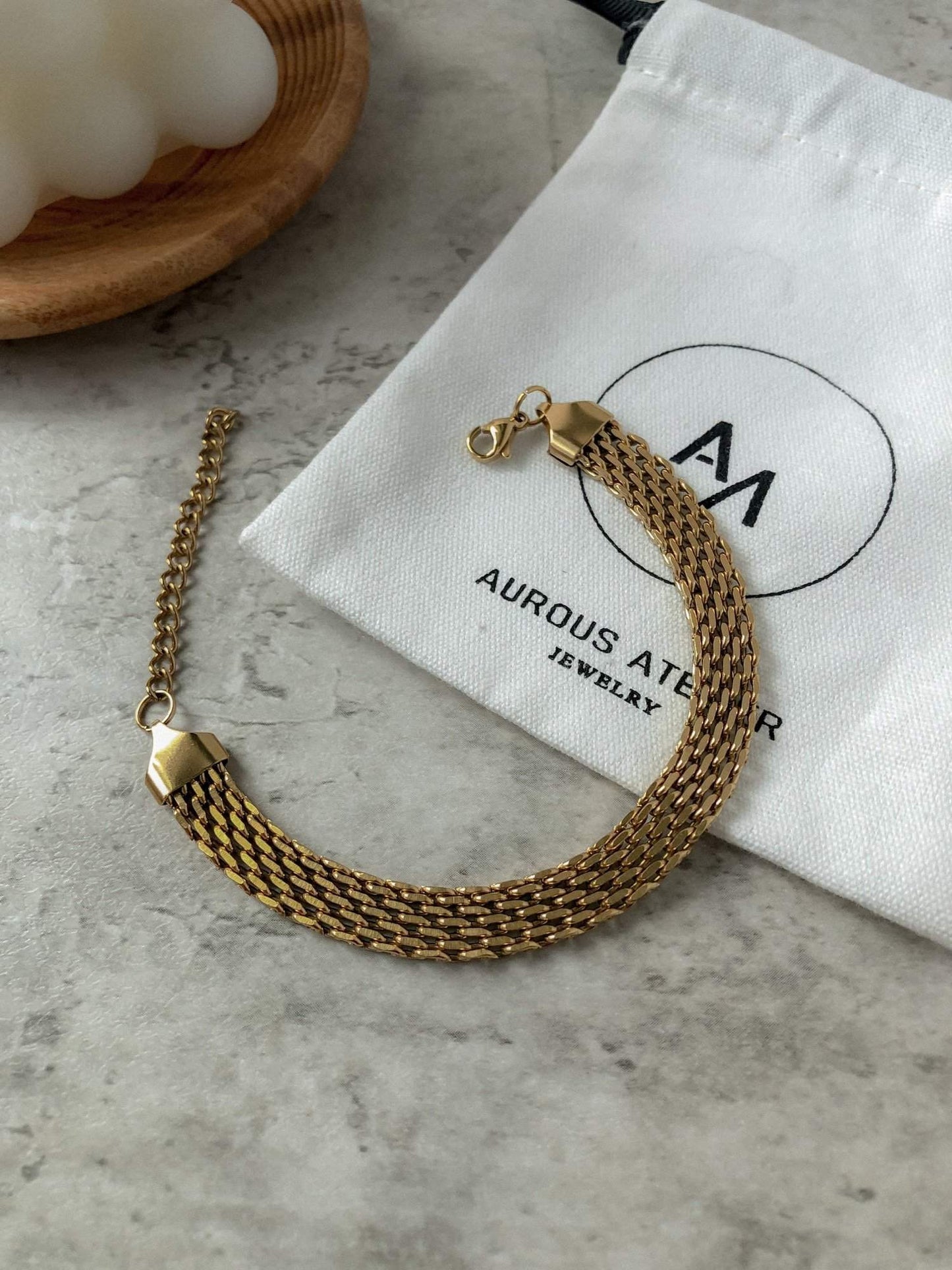 A clean studio macro layout showing the flat, continuous row pattern of the Danielle gold mesh chain bracelet next to its organic canvas canvas brand pouch.