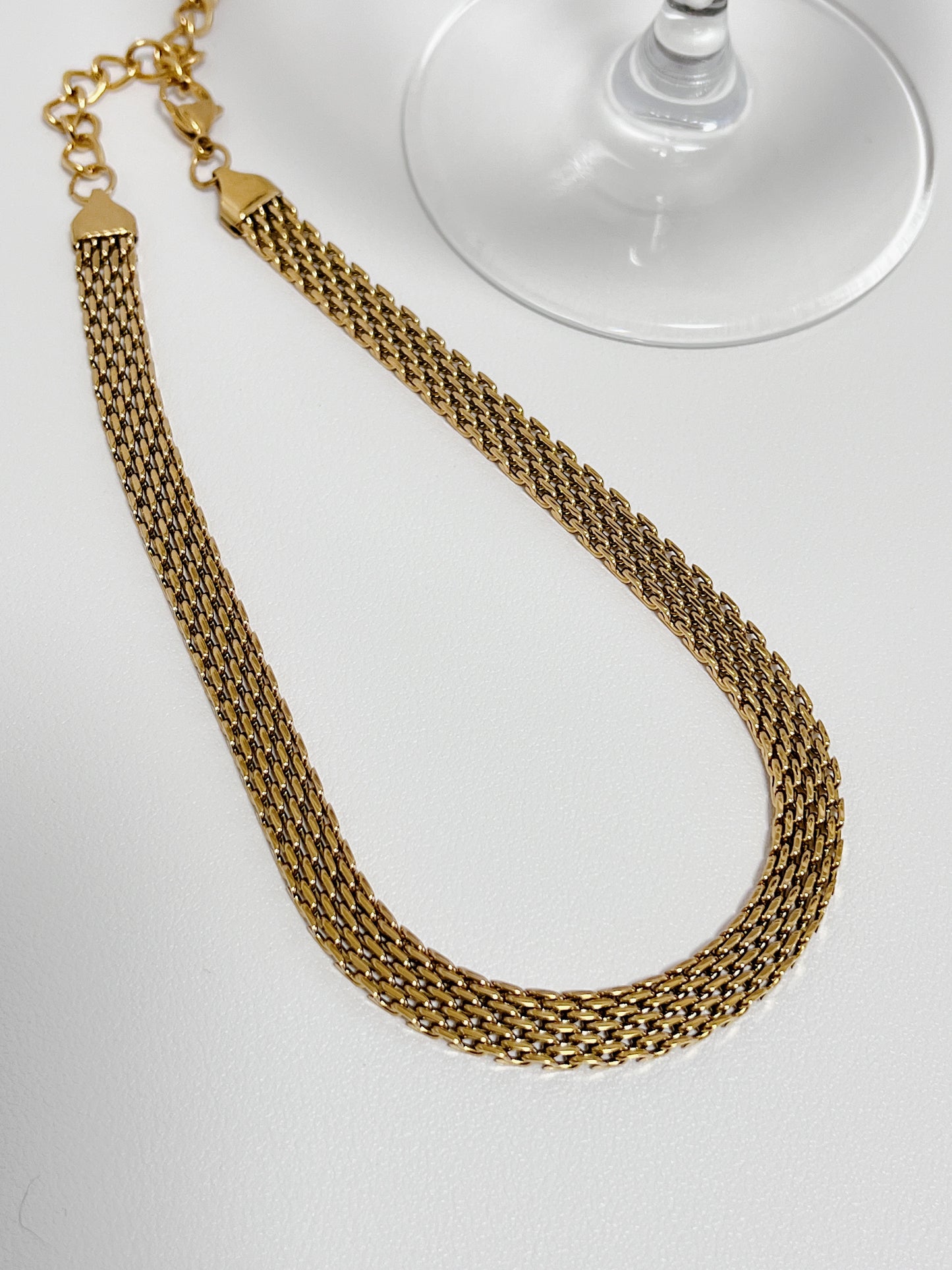 Studio flat lay of the Danielle mesh chain necklace showing its wide 8mm gold profile next to a clear glass stem