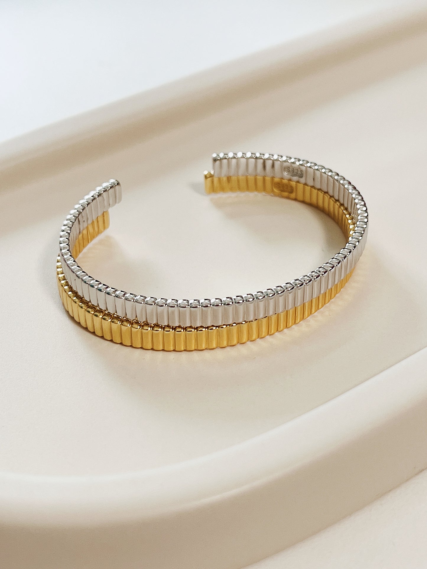 Parallel alignment of mixed-metal demi-fine jewelry stacking bangles resting horizontally on a minimalist neutral tray background.