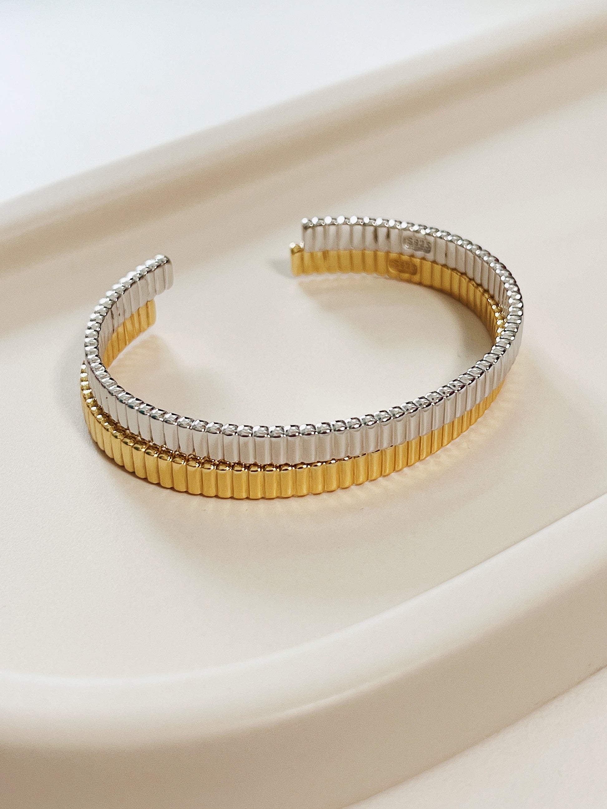Parallel alignment of mixed-metal demi-fine jewelry stacking bangles resting horizontally on a minimalist neutral tray background.