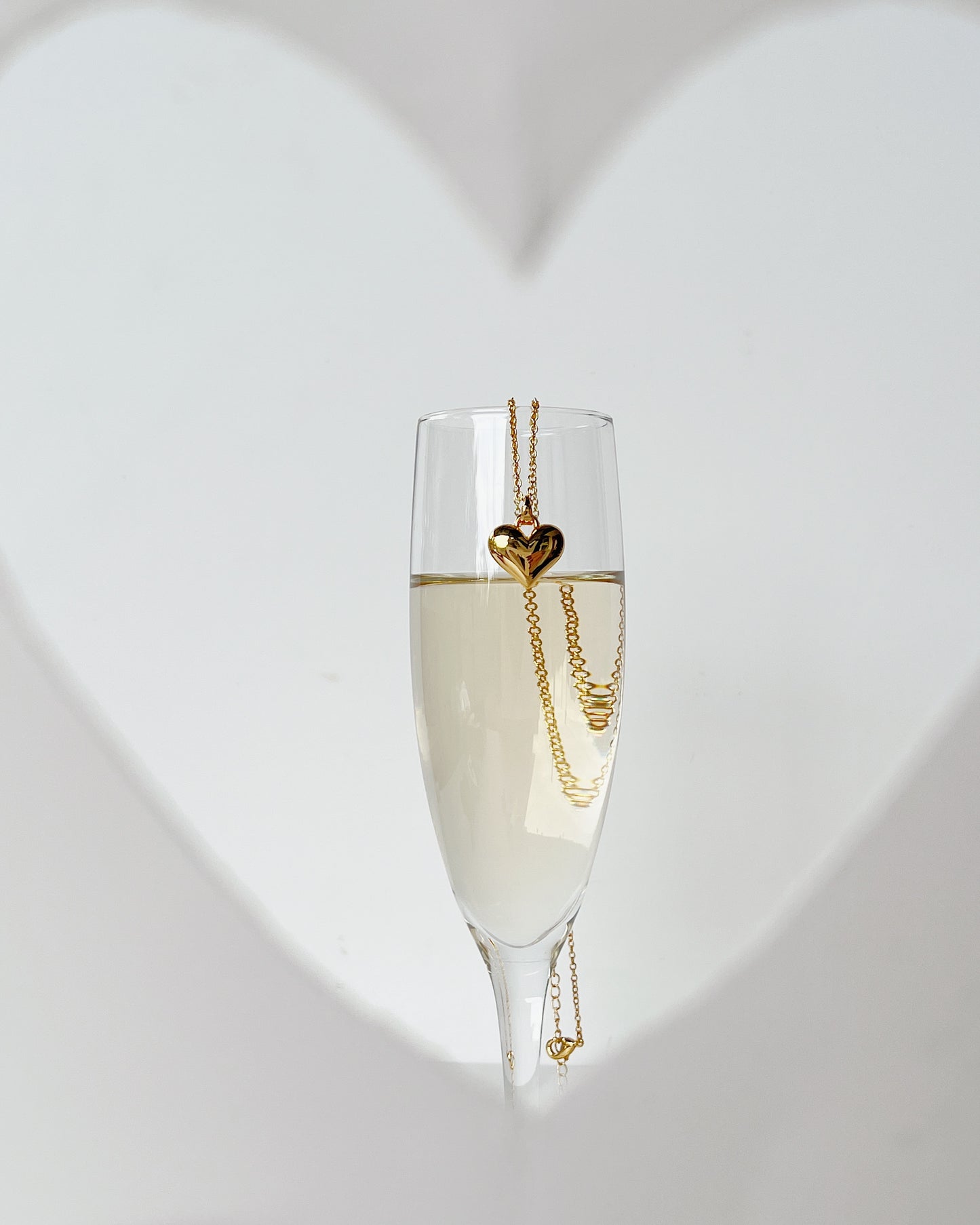 An editorial studio concept showing the solid gold Lia heart pendant hanging on the rim of a filled champagne flute against a blurred white heart background shape