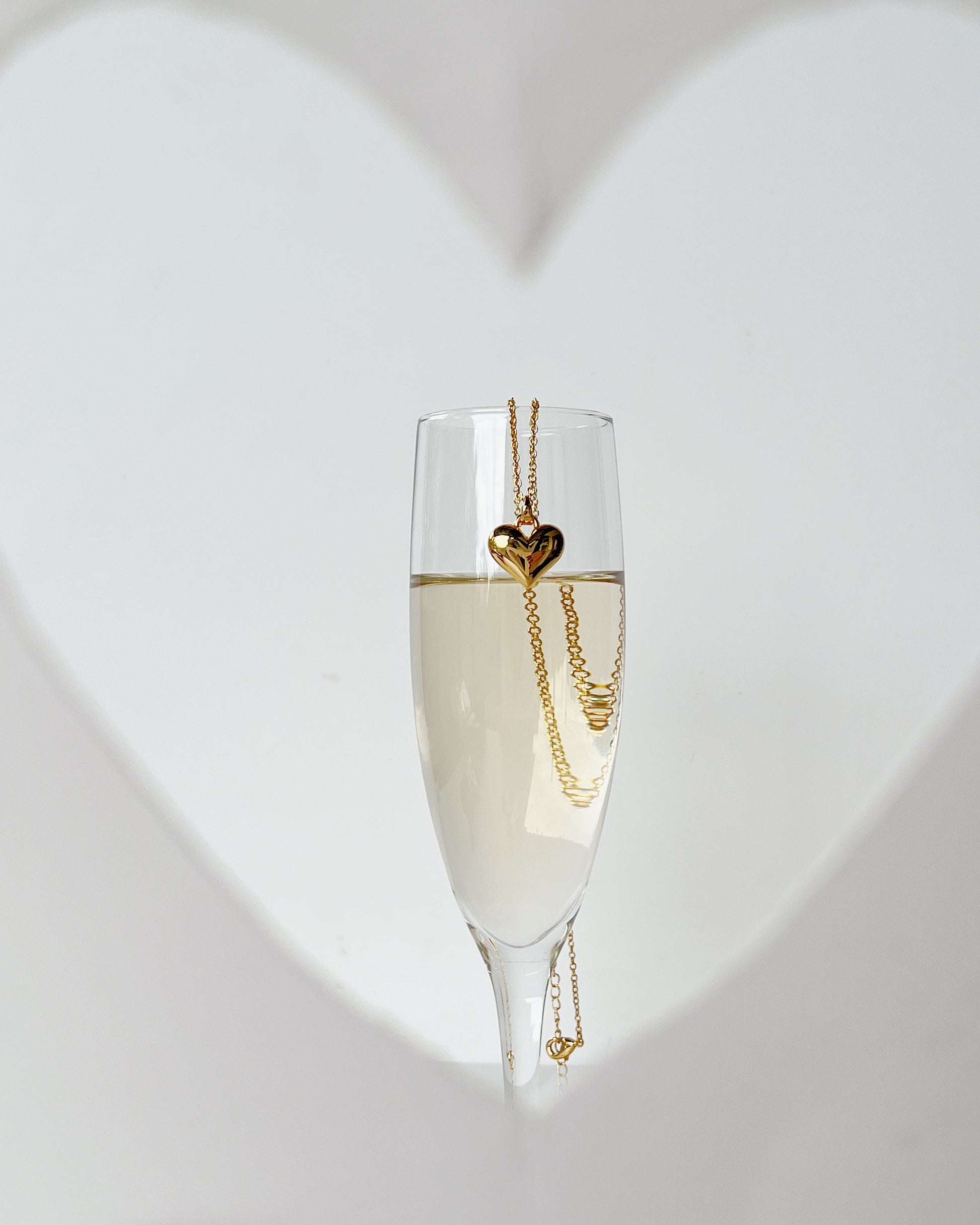 An editorial studio concept showing the solid gold Lia heart pendant hanging on the rim of a filled champagne flute against a blurred white heart background shape