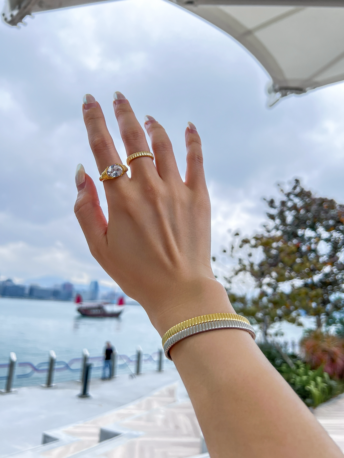 Editorial lookbook shot of a model's hand raised toward the Hong Kong harbor skyline, wearing the gold and silver flexi cuff stack paired with bold statement rings.
