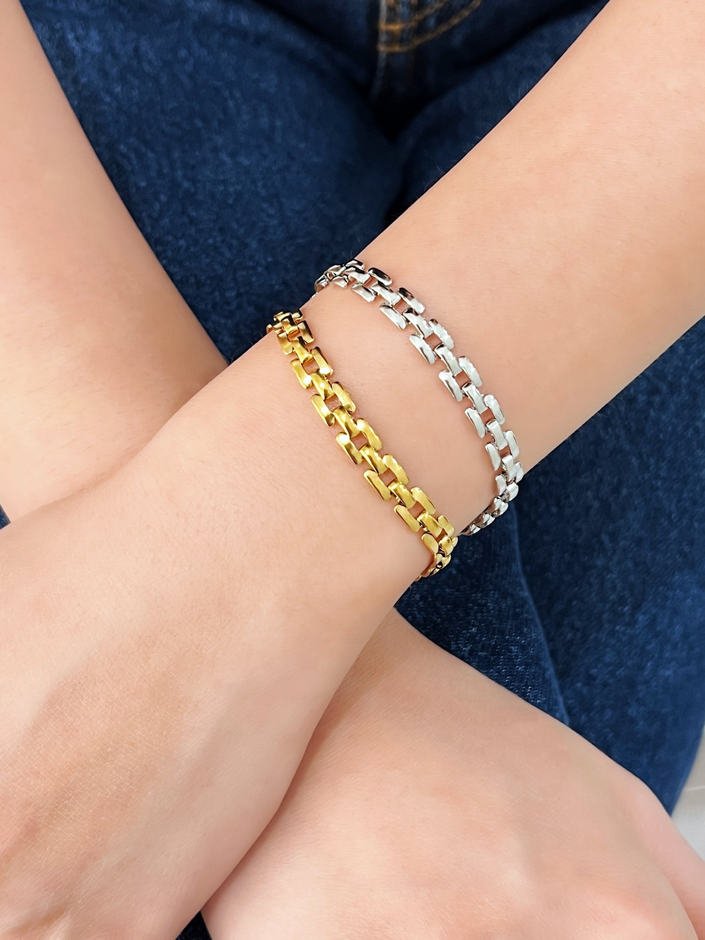 Close-up shot of a model’s crossed wrists styling the gold and silver Evie link bracelets together over dark denim pants.