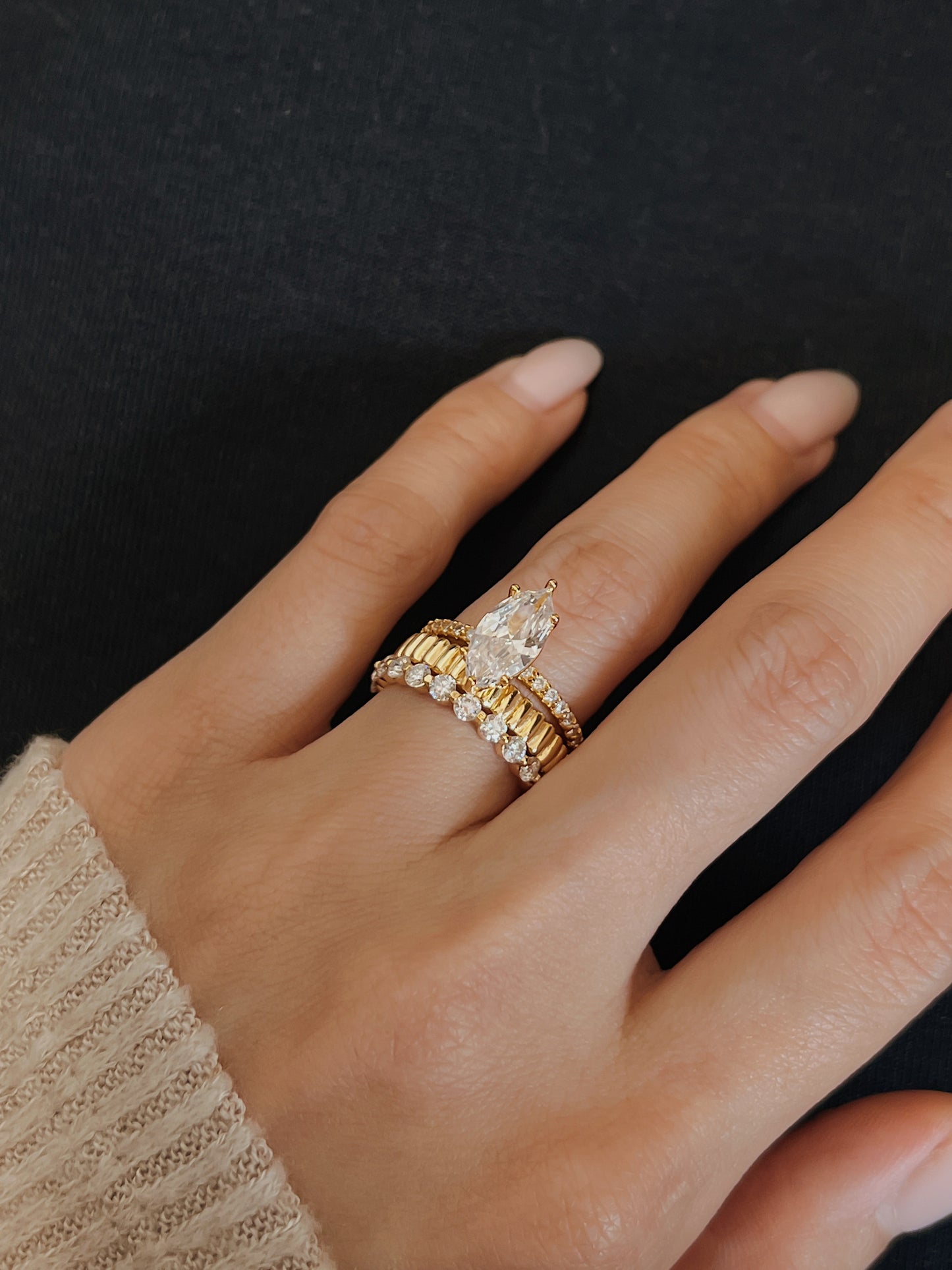 Close up lifestyle shot of a floating shared prong moissanite eternity band styled with a vede ribbed gold ring and a cream ribbed sweater cuff