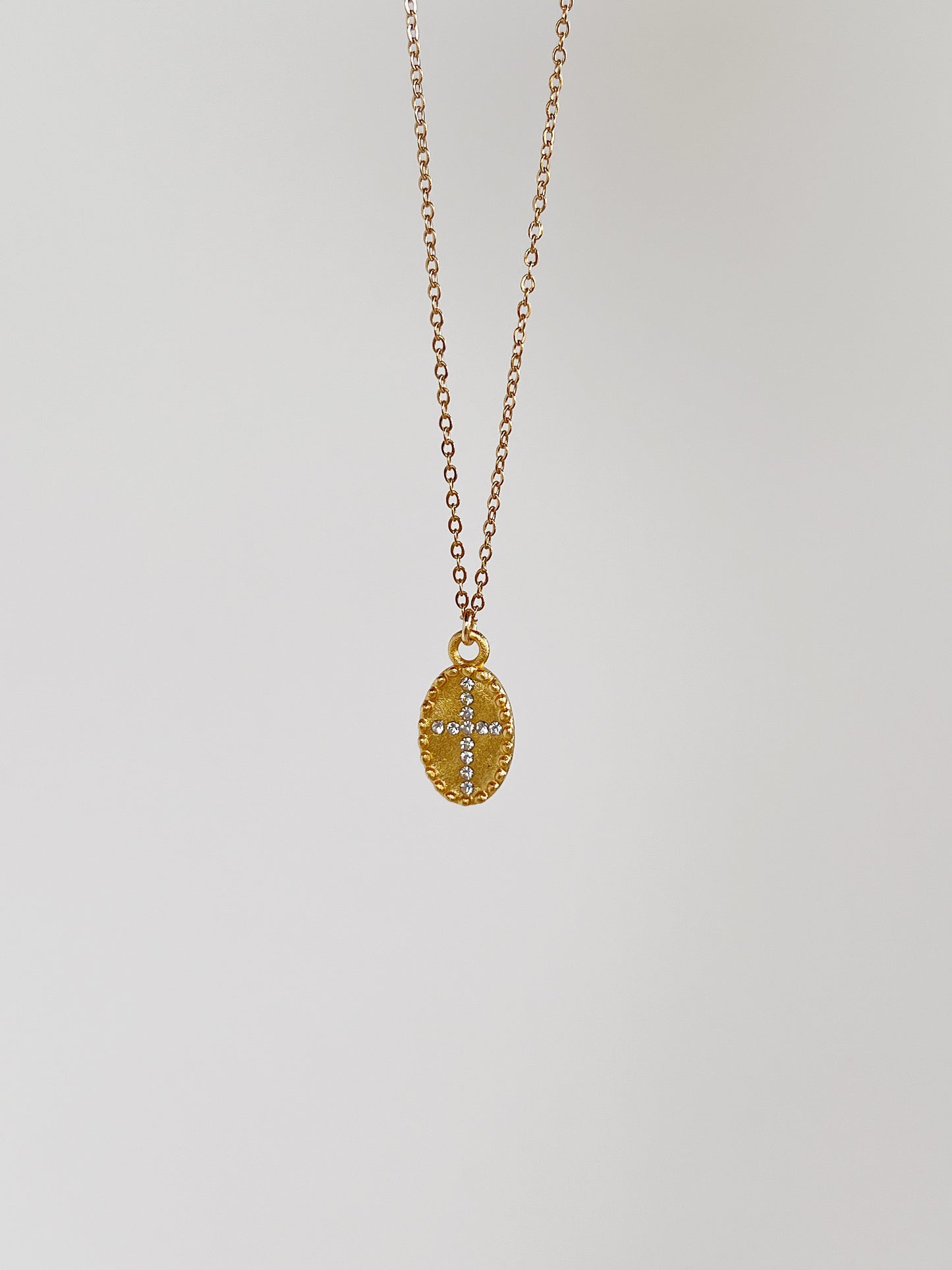 The gold Gianna oval cross medallion necklace suspended vertically against a minimalist, clean white studio background to showcase the fluid chain drape.