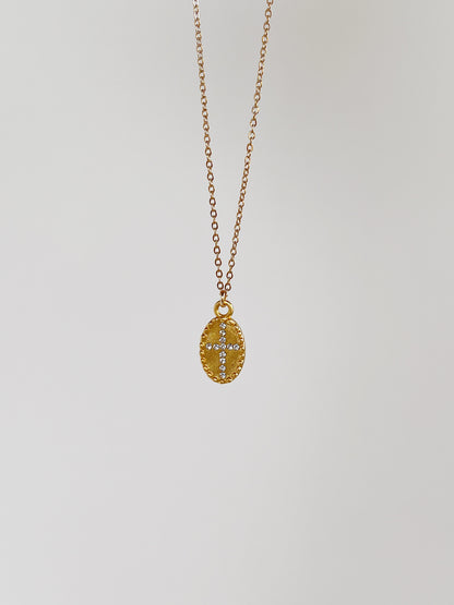 The gold Gianna oval cross medallion necklace suspended vertically against a minimalist, clean white studio background to showcase the fluid chain drape.