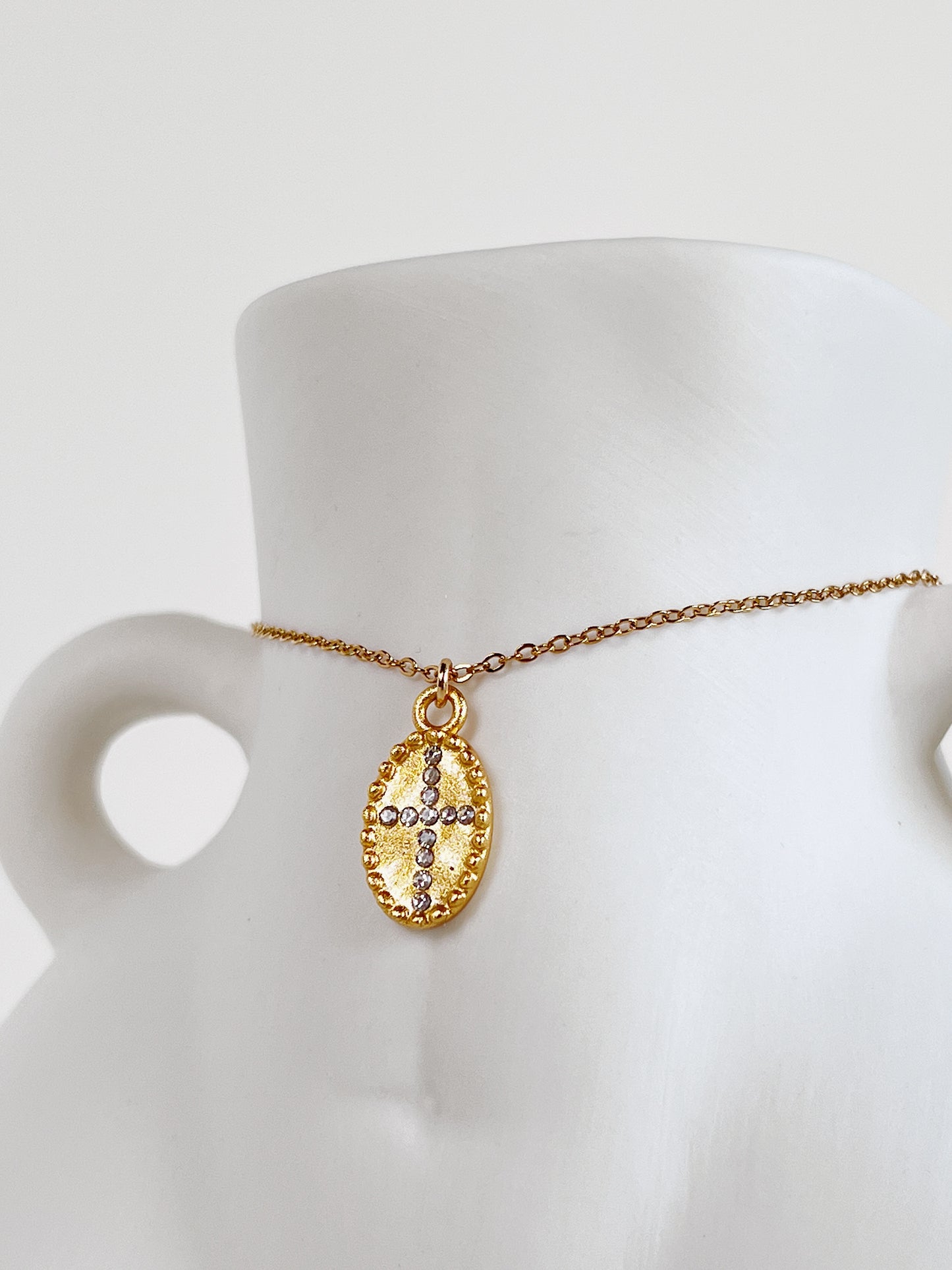 The Gianna 18k gold PVD oval cross medallion necklace draped over the waist of a minimalist white ceramic female body-sculpture vase