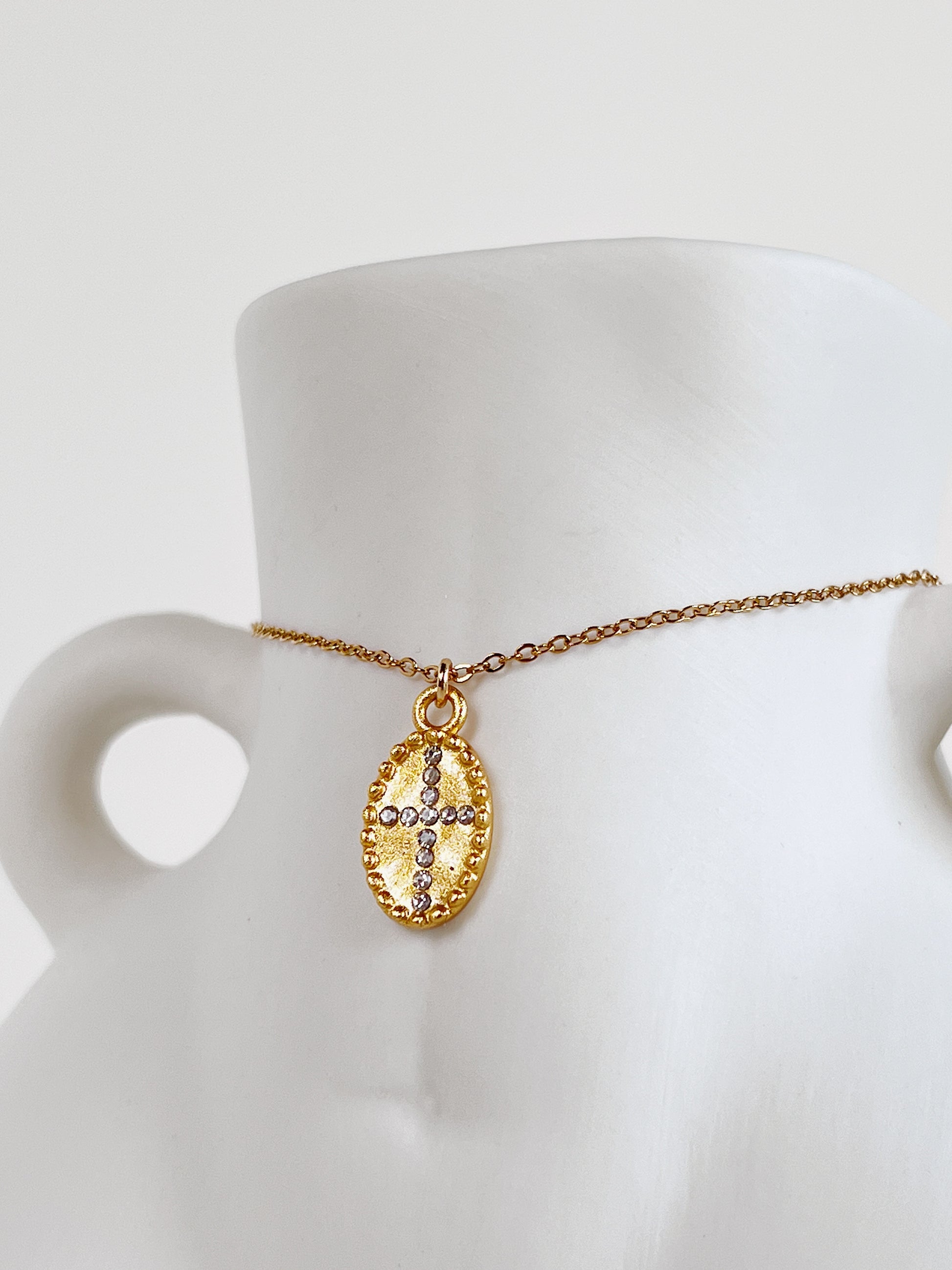 The Gianna 18k gold PVD oval cross medallion necklace draped over the waist of a minimalist white ceramic female body-sculpture vase