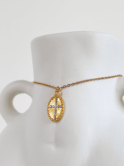 The Gianna 18k gold PVD oval cross medallion necklace draped over the waist of a minimalist white ceramic female body-sculpture vase