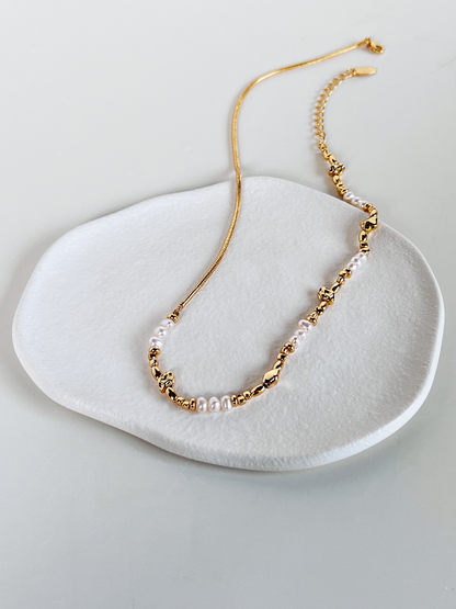 An overhead, elevated view of the Greta asymmetric necklace resting on a white, organic-shaped ceramic display dish. The image clearly shows the contrast between the fluid gold snake chain and the textured strand of natural freshwater pearls and sculptural molten gold beads.