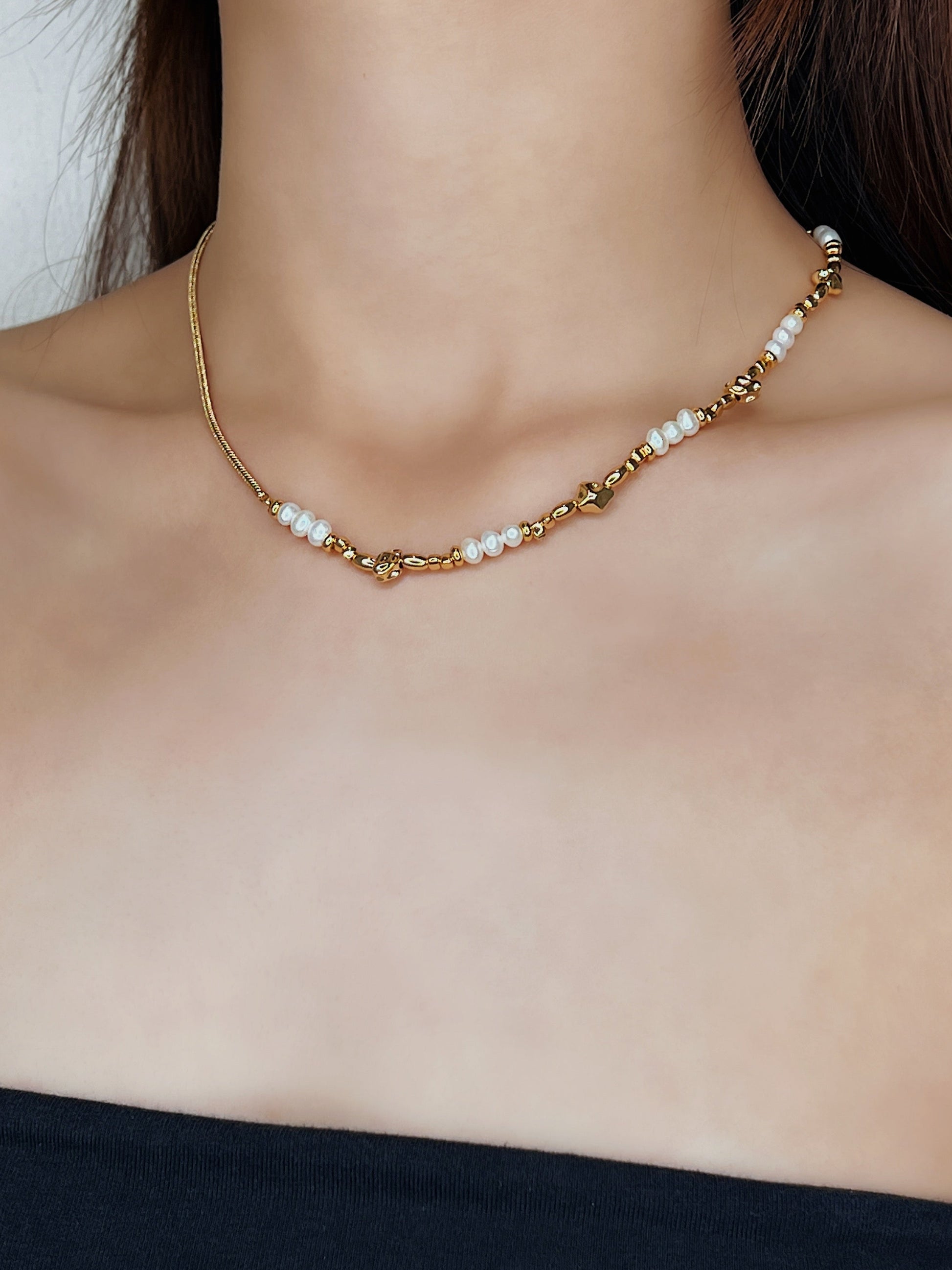A side-angled portrait of a model wearing the Greta asymmetric necklace. The shot beautifully illustrates how the minimalist gold chain sits on one side of the neck, while the strand of organic freshwater pearls and molten gold beads sits on the other, creating a unique visual balance.