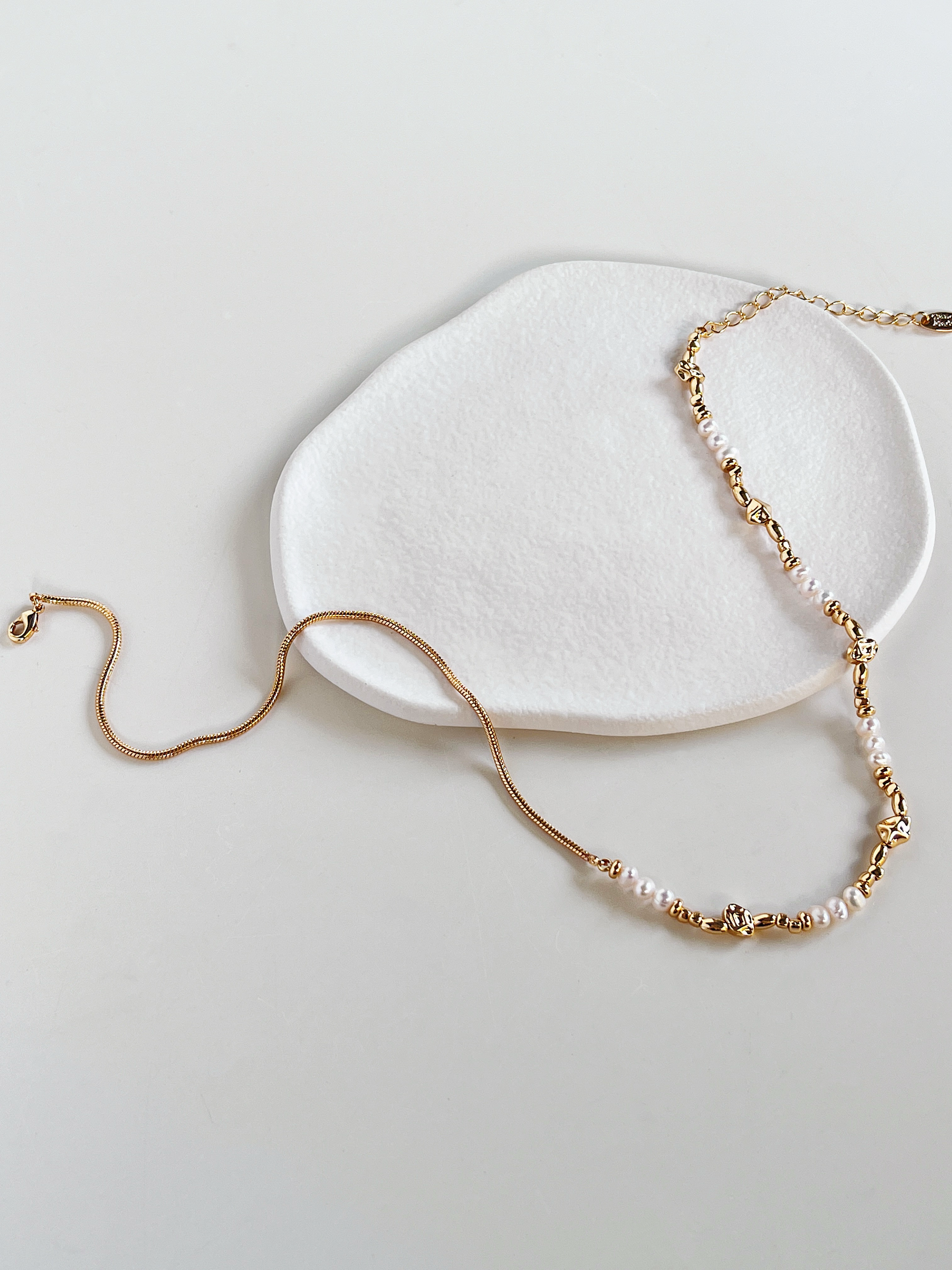 A wide, open laydown of the Greta asymmetric necklace, showcasing the winding, fluid quality of the gold snake chain contrasting with the structured baroque pearls and irregular molten gold beads on a white background.