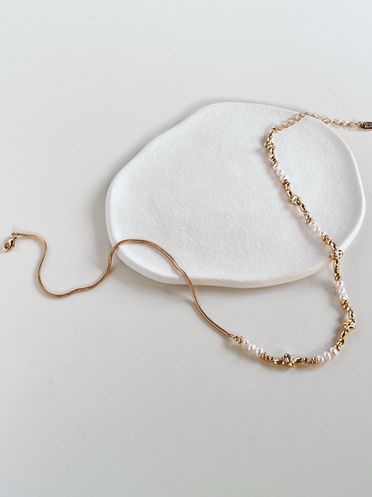 A wide, open laydown of the Greta asymmetric necklace, showcasing the winding, fluid quality of the gold snake chain contrasting with the structured baroque pearls and irregular molten gold beads on a white background.