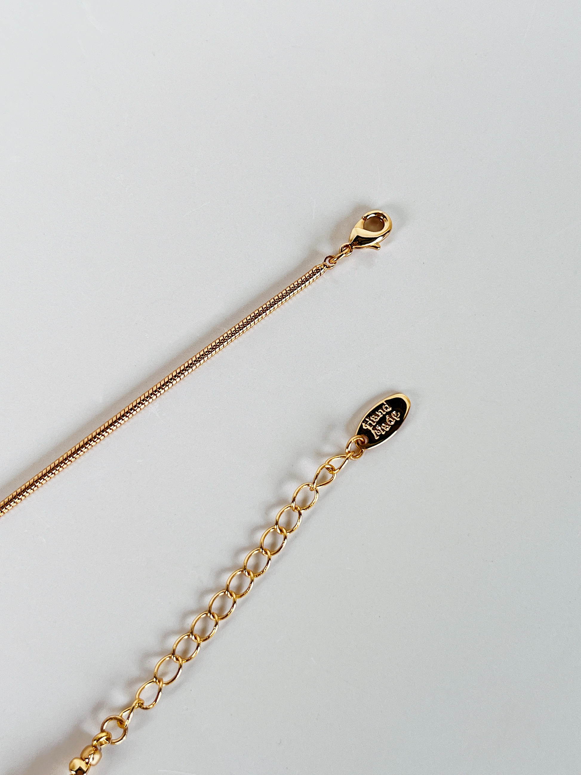 A high-resolution close-up of the Greta necklace's closure system, featuring a polished gold lobster clasp, a delicate extender chain, and a distinct oval gold tag engraved with the word "Handmade." This shot emphasizes the artisan quality of the piece.