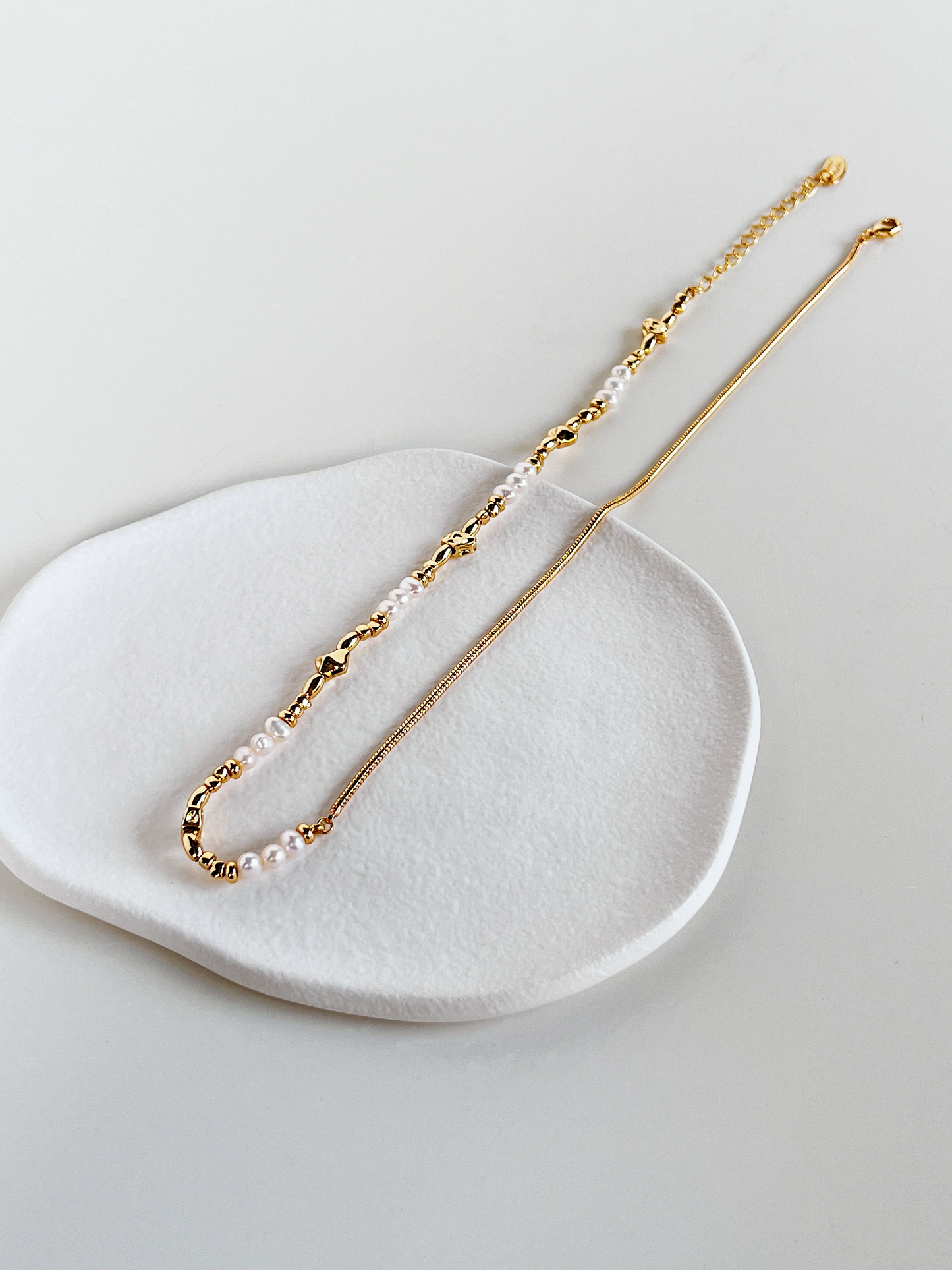 A side-angled laydown view of the Greta asymmetric pearl and gold chain necklace on a white ceramic dish. The image captures the full length of the piece, highlighting the modern blend of textures from a unique perspective.
