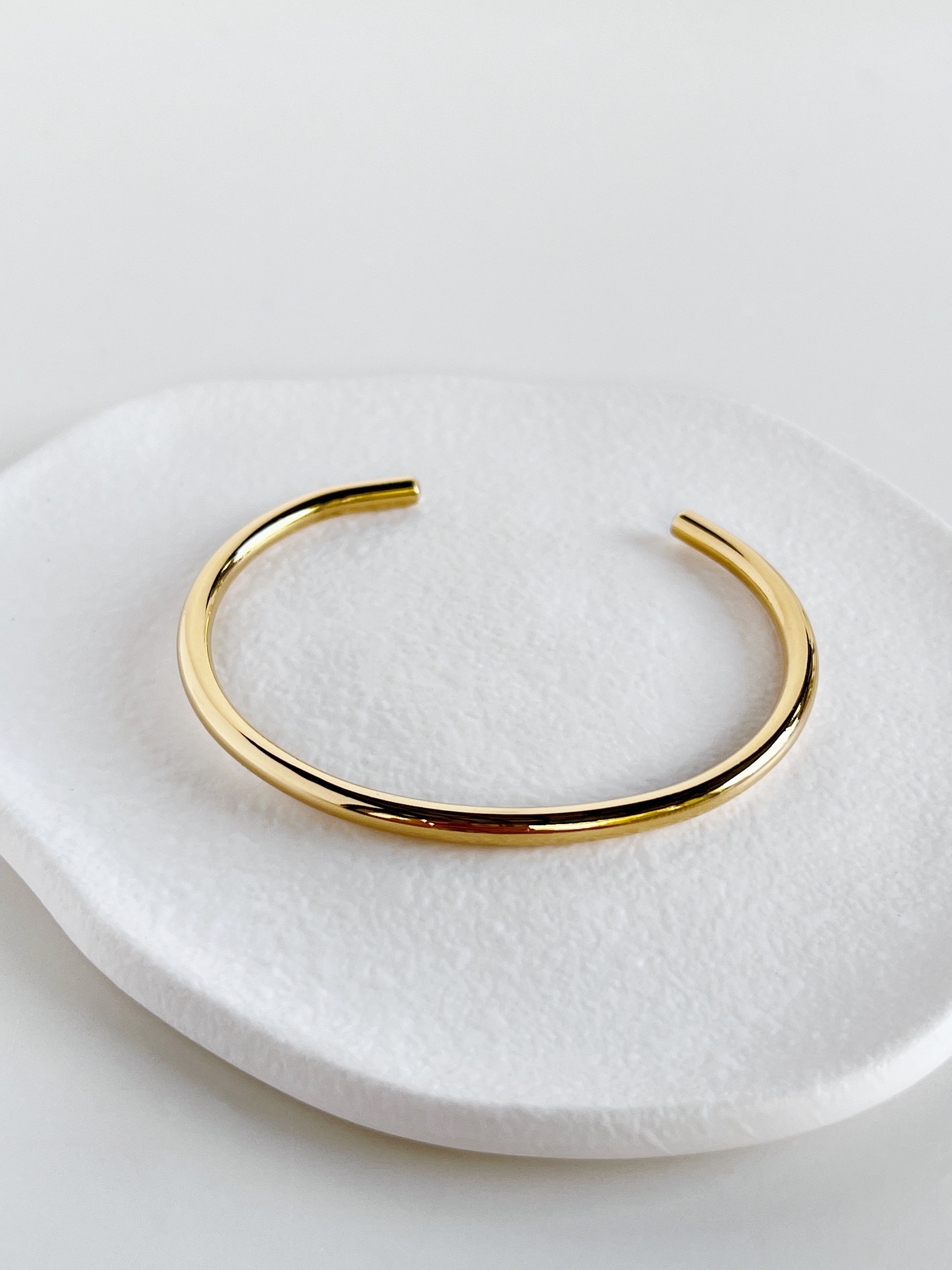 High-angle macro shot of the Ines minimal cuff bangle resting on an organic white textured stone plate, showcasing the high-density 18K gold mirror finish.