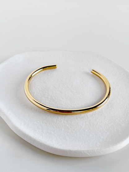 High-angle macro shot of the Ines minimal cuff bangle resting on an organic white textured stone plate, showcasing the high-density 18K gold mirror finish.
