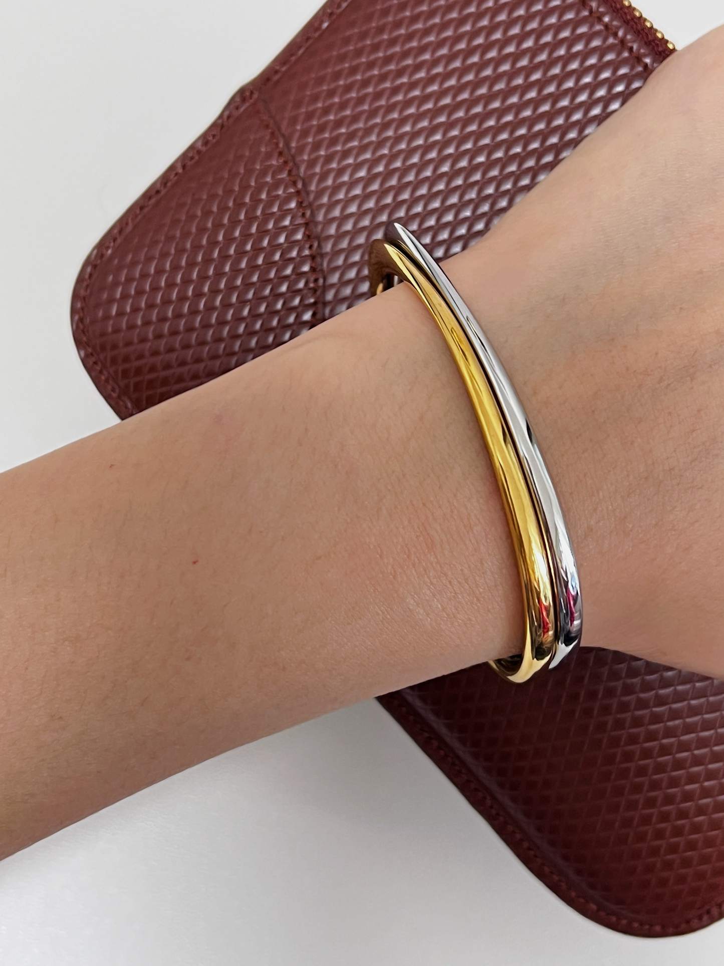 A high-angle view of a model's wrist wearing the two-tone gold and silver Iva undulating cuffs stacked elegantly over a textured rich brown leather clutch bag
