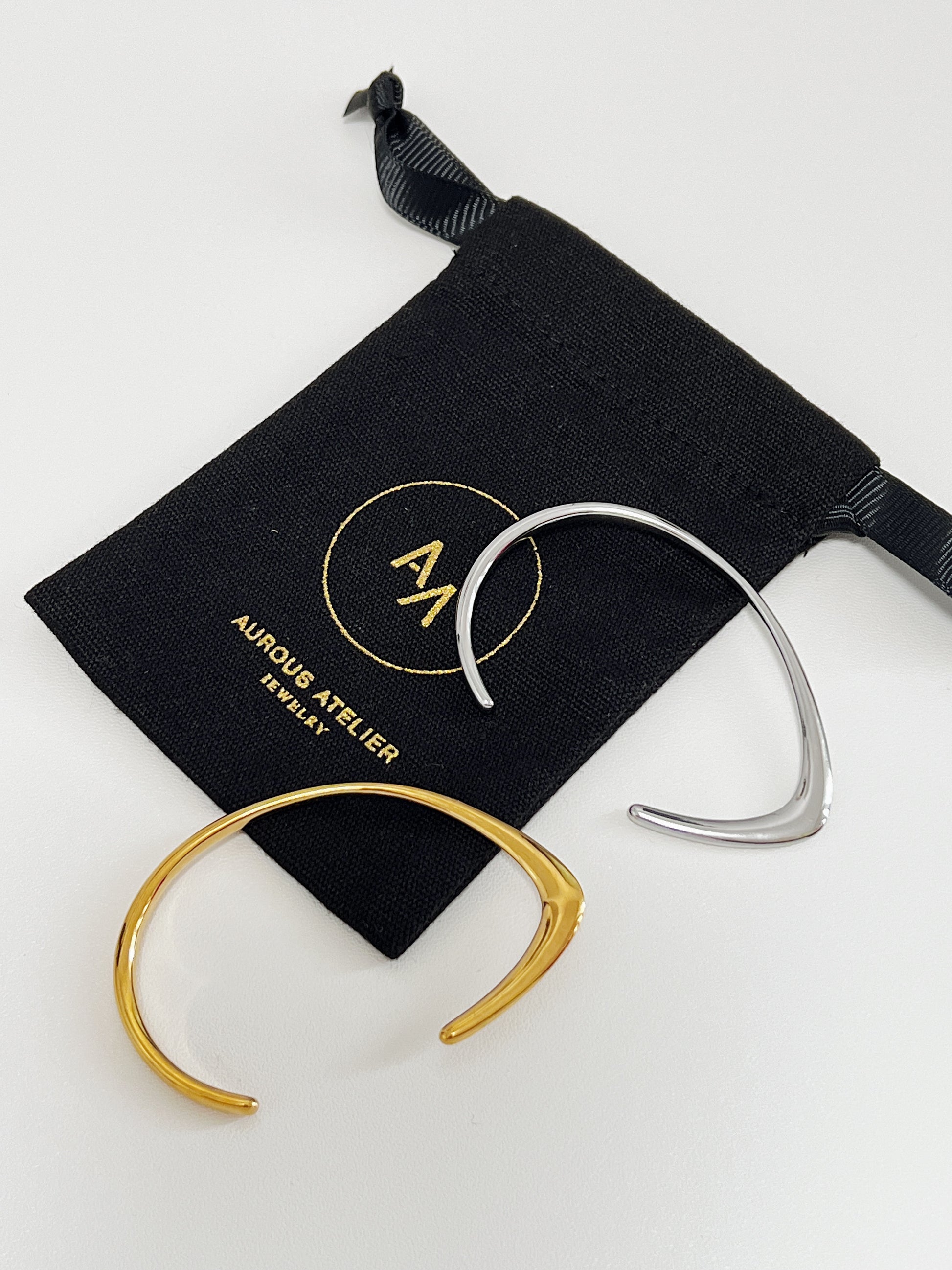 A premium product shot displaying the gold and silver variants of the Iva irregular open-cuff bangle resting gracefully on a black velvet Aurous Atelier jewelry pouch, highlighting their unique transition from a sharp knife-edge to a smooth rounded curve