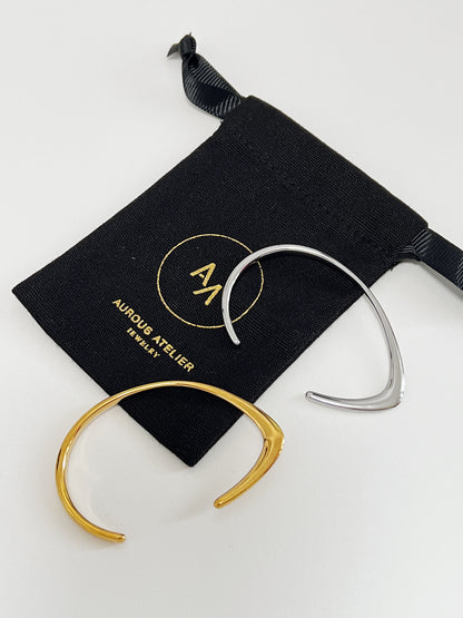 A premium product shot displaying the gold and silver variants of the Iva irregular open-cuff bangle resting gracefully on a black velvet Aurous Atelier jewelry pouch, highlighting their unique transition from a sharp knife-edge to a smooth rounded curve