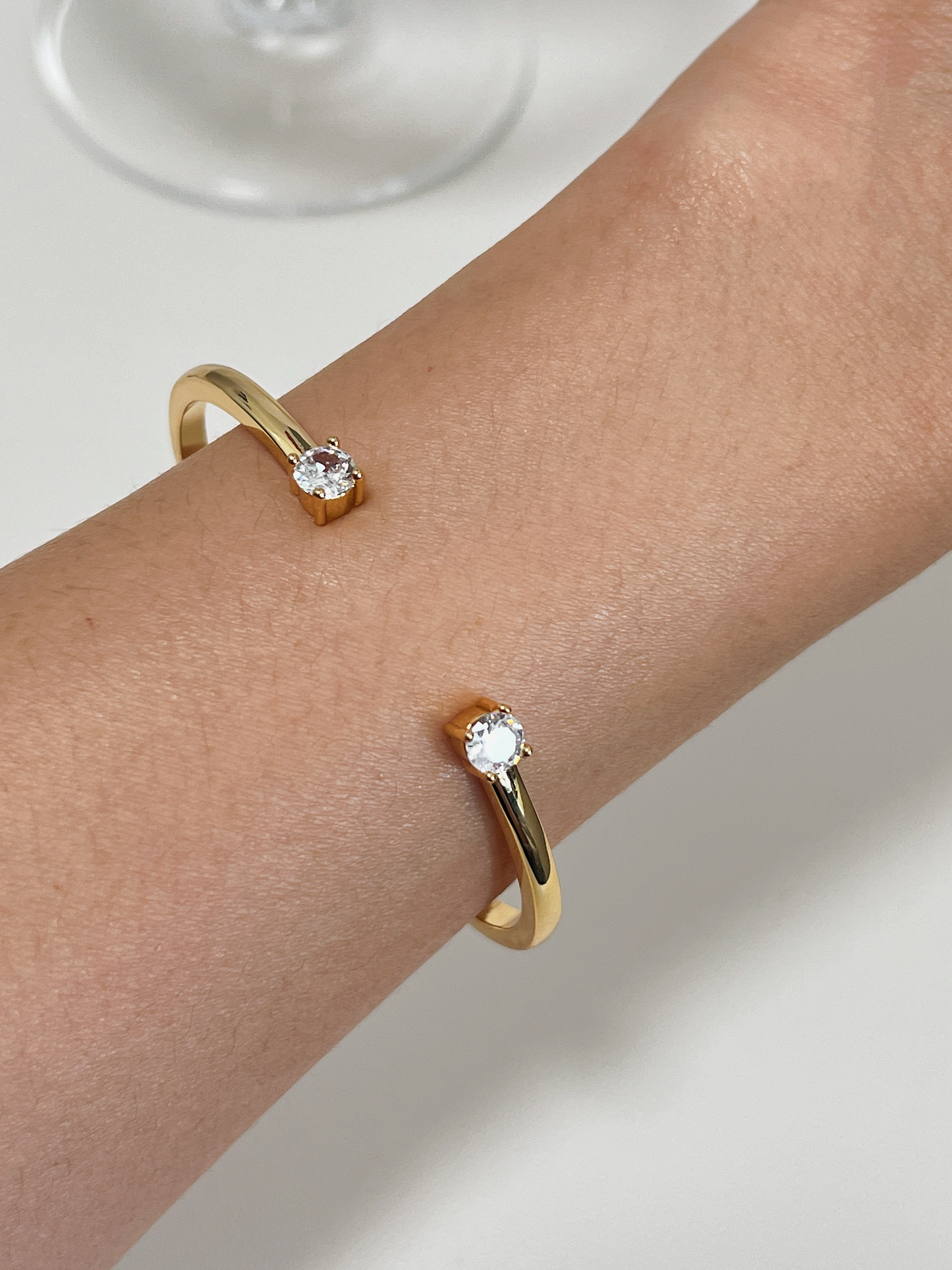 Close-up of a model's wrist wearing the sleek Justine gold open-bangle bracelet while resting her hand near a delicate wine glass.