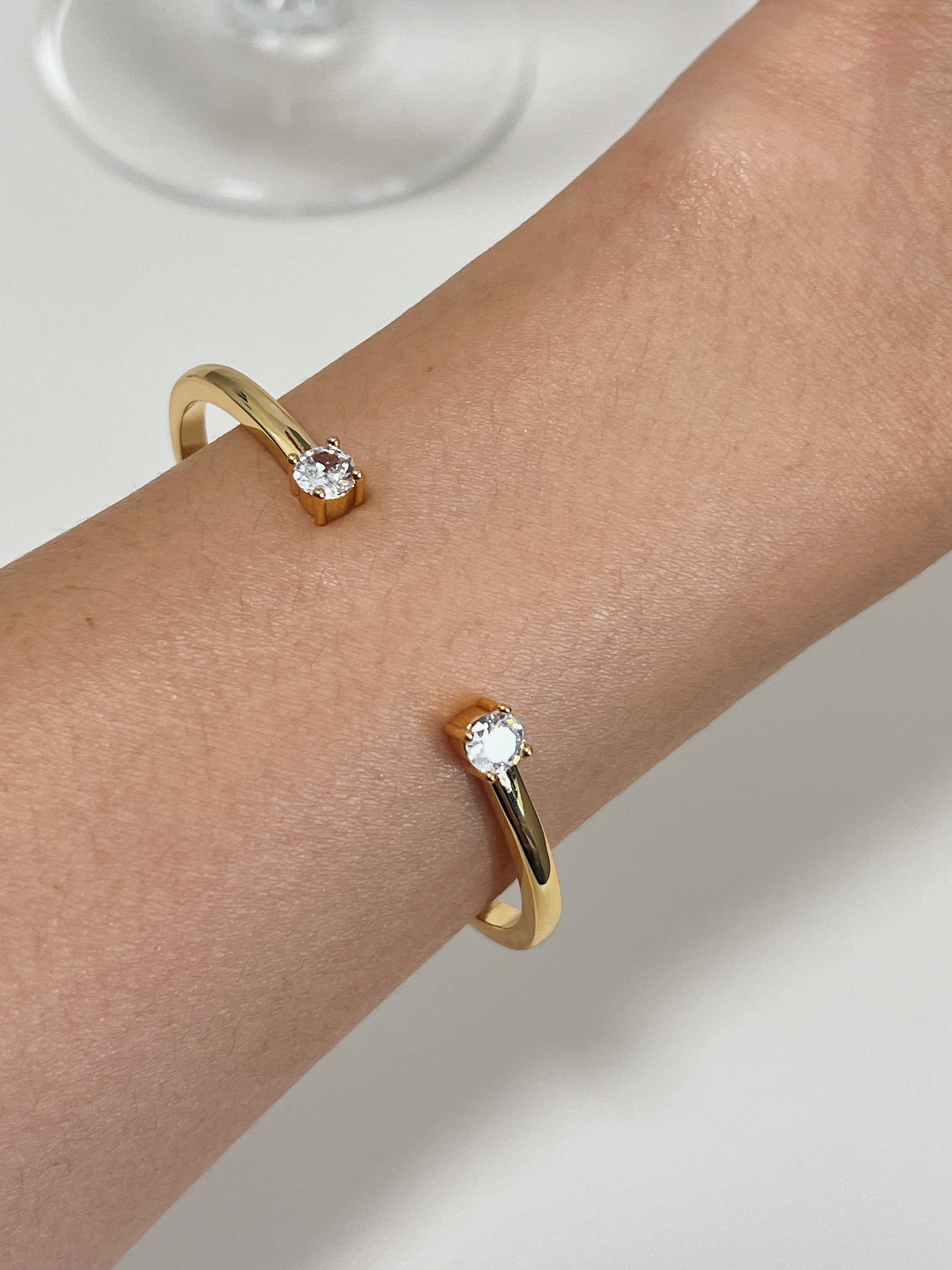 Close-up of a model's wrist wearing the sleek Justine gold open-bangle bracelet while resting her hand near a delicate wine glass.