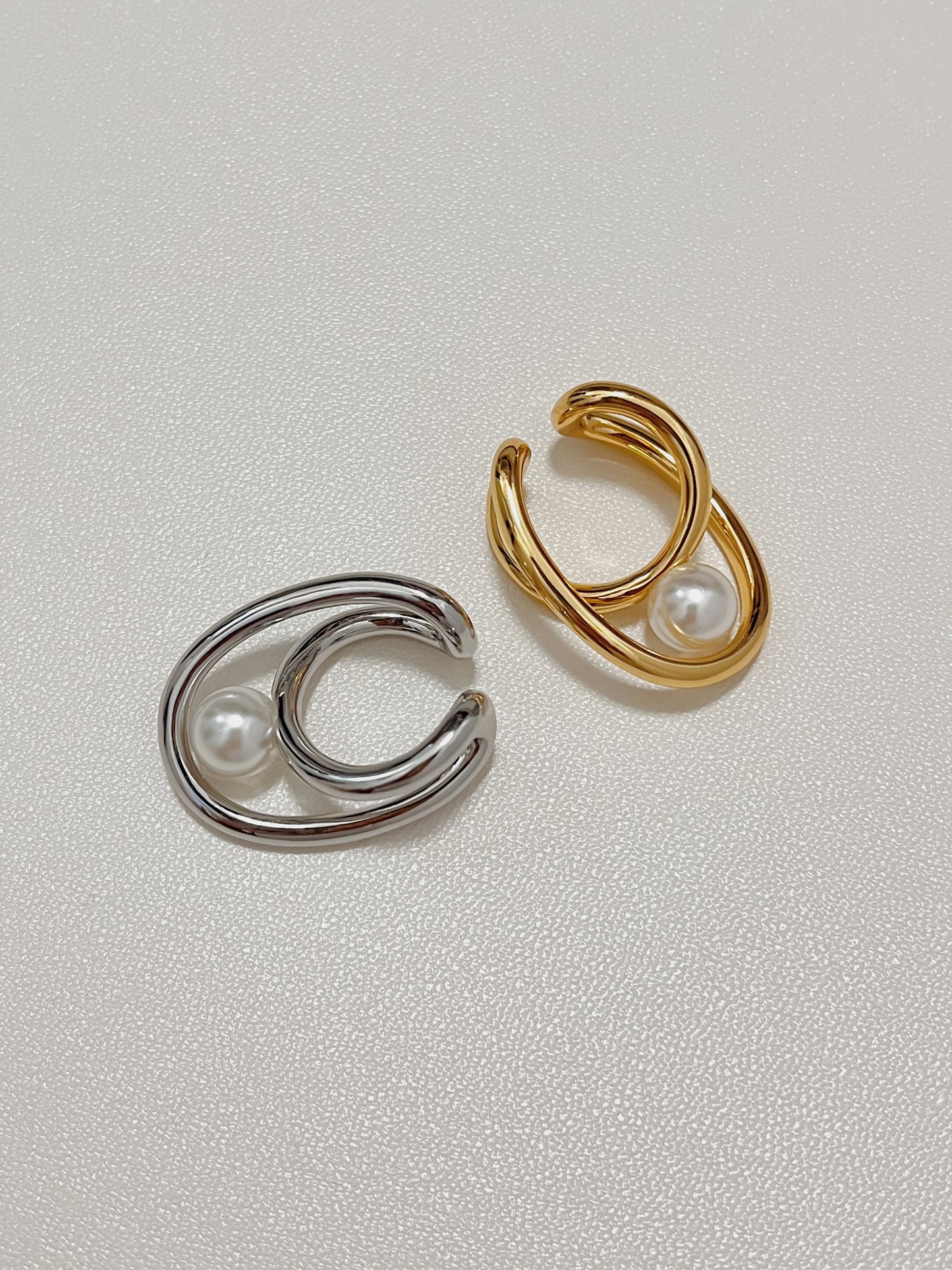 Top-down product shot featuring the Karolina Statement Pearl Earlobe Cuff in both a high-shine silver and a luxurious warm gold finish, showcasing their nested sculptural loops and central glowing pearls on a textured white background.