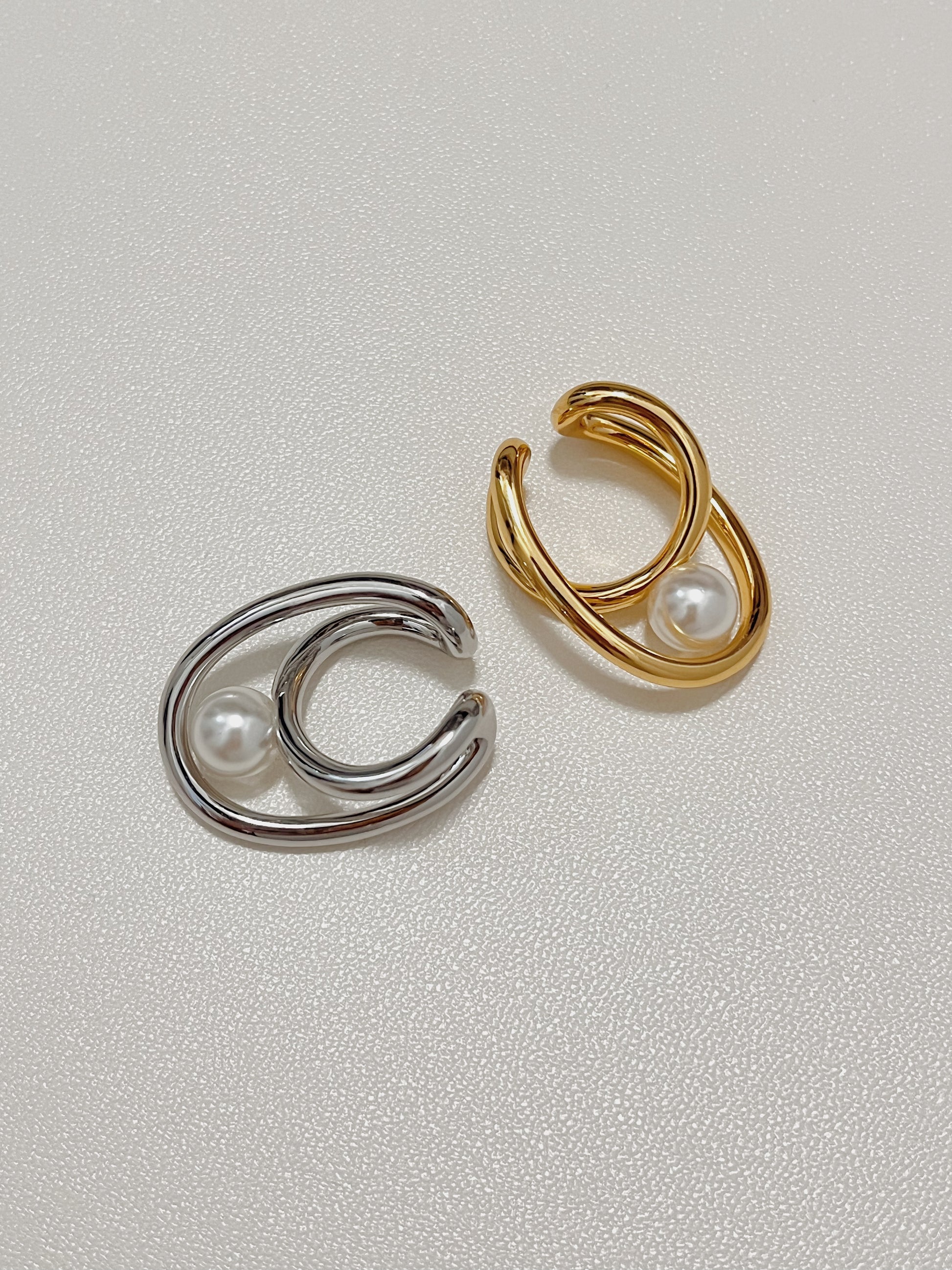 Top-down product shot featuring the Karolina Statement Pearl Earlobe Cuff in both a high-shine silver and a luxurious warm gold finish, showcasing their nested sculptural loops and central glowing pearls on a textured white background.