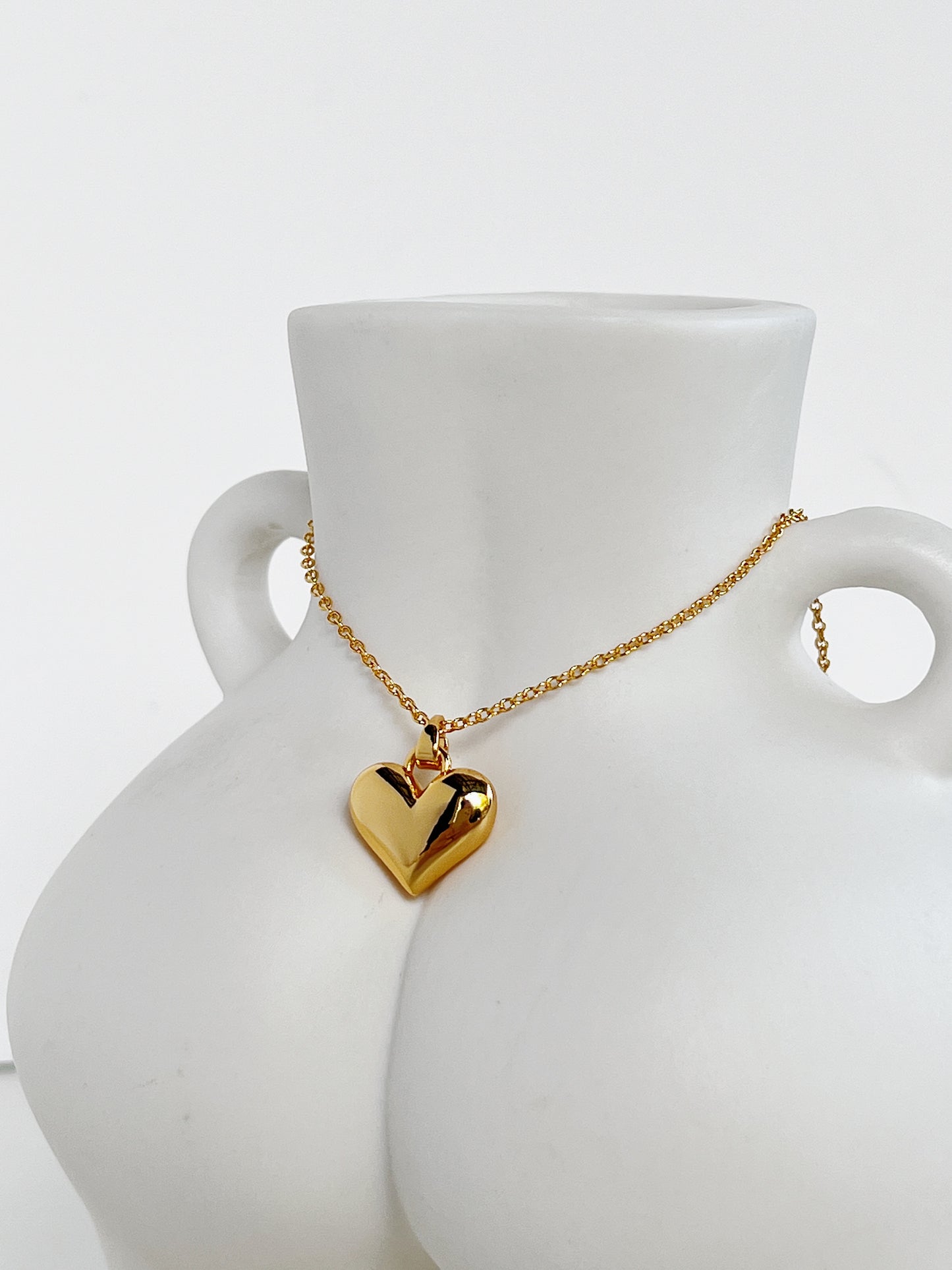 A studio view of the solid gold Lia heart necklace draped elegantly across a minimalist white ceramic body-sculpture vase