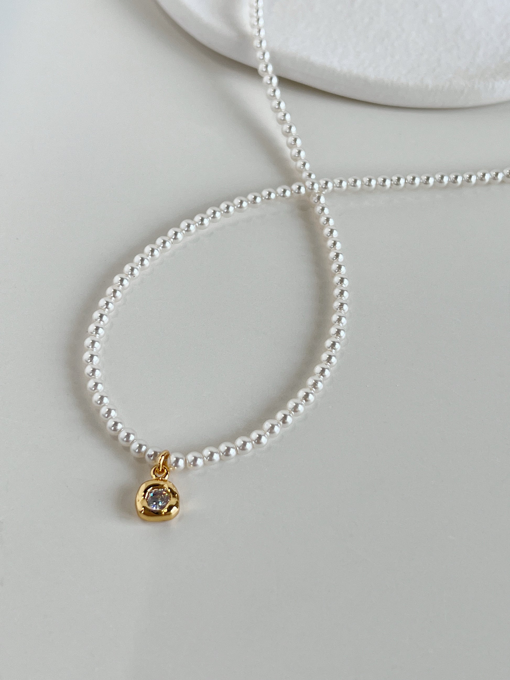A high-end catalog flatlay of the Alta pearl necklace displaying the uniform 4mm white seed pearls and its central organic gold bezel charm on a white background