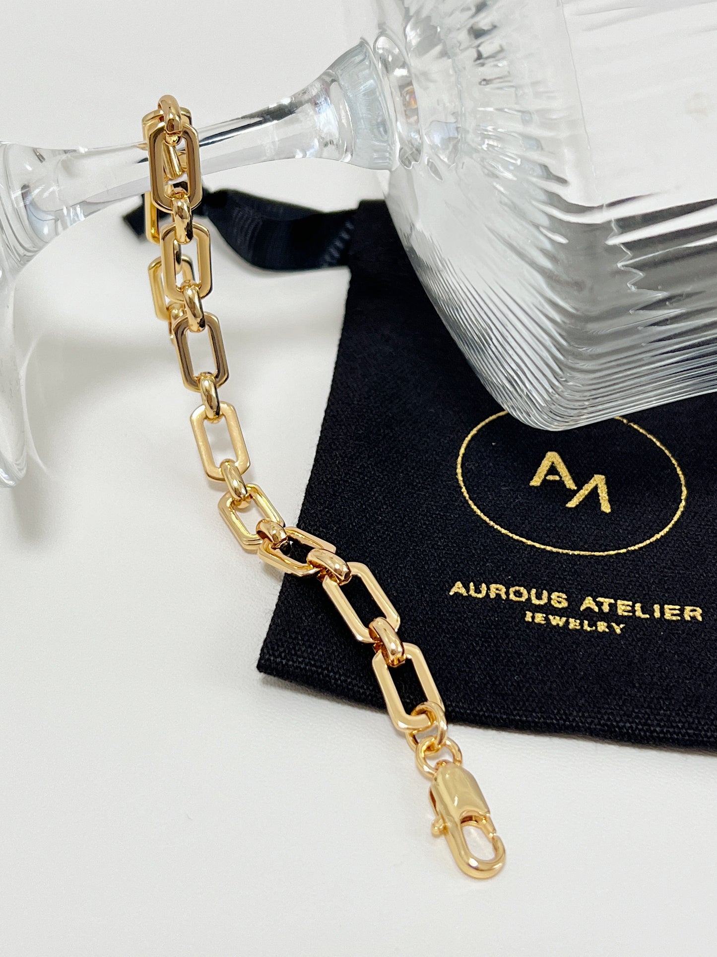 The 7mm gold link bracelet draped elegantly under the clear stem of a crystal glass, resting across a signed black Aurous Atelier jewelry pouch.