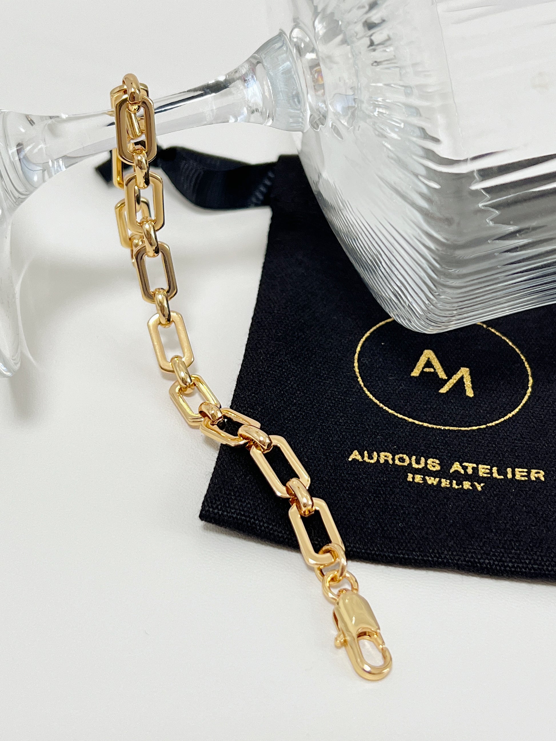 The 7mm gold link bracelet draped elegantly under the clear stem of a crystal glass, resting across a signed black Aurous Atelier jewelry pouch.