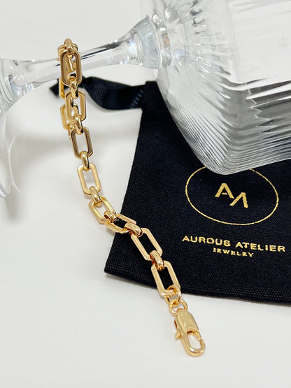 The 7mm gold link bracelet draped elegantly under the clear stem of a crystal glass, resting across a signed black Aurous Atelier jewelry pouch.