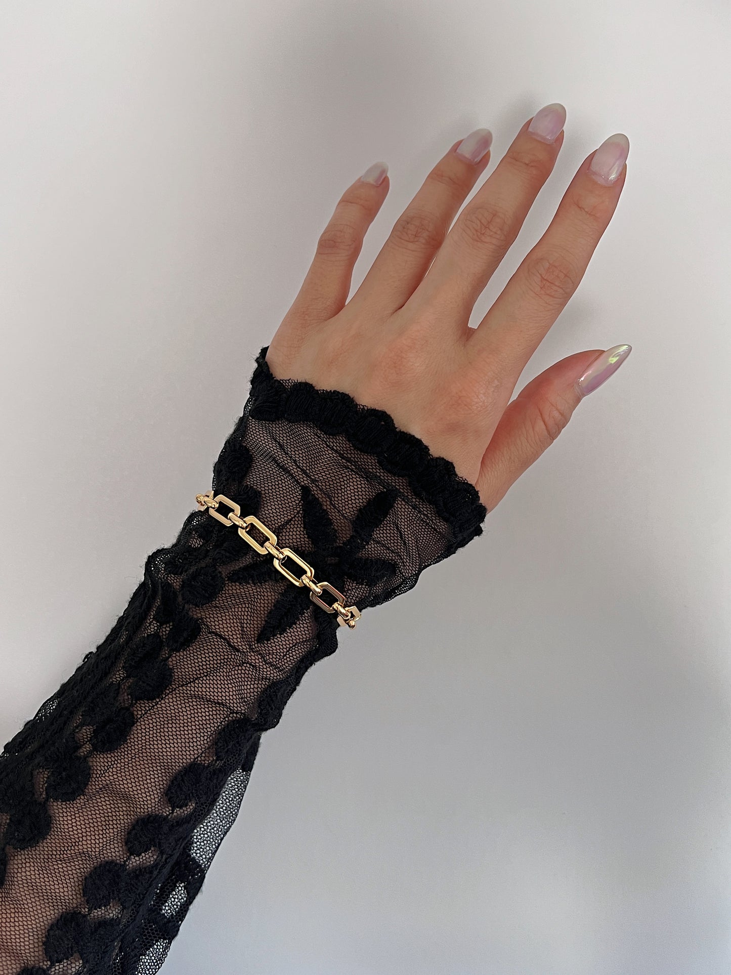 Macro detail shot of the rectangular beveled links of the Maia gold chain catching the light over a dramatic black lace cuff garment.