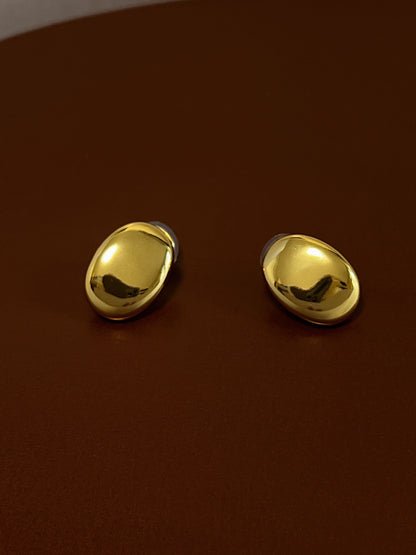 High-polish pair of sleek, gold oval clip on earrings resting on a rich brown leather background.