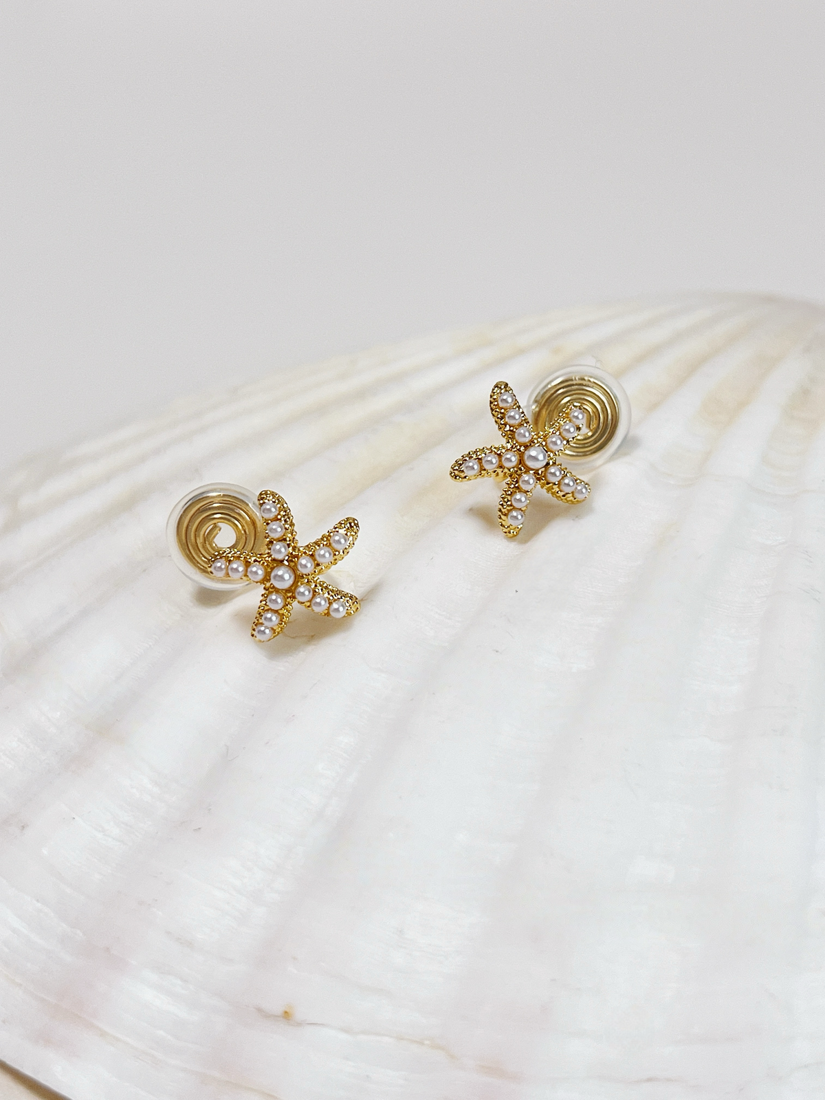 Pair of pearl starfish clip-ons displayed on a white seashell prop, showing the golden spiral clip backs and silicone cushioning.